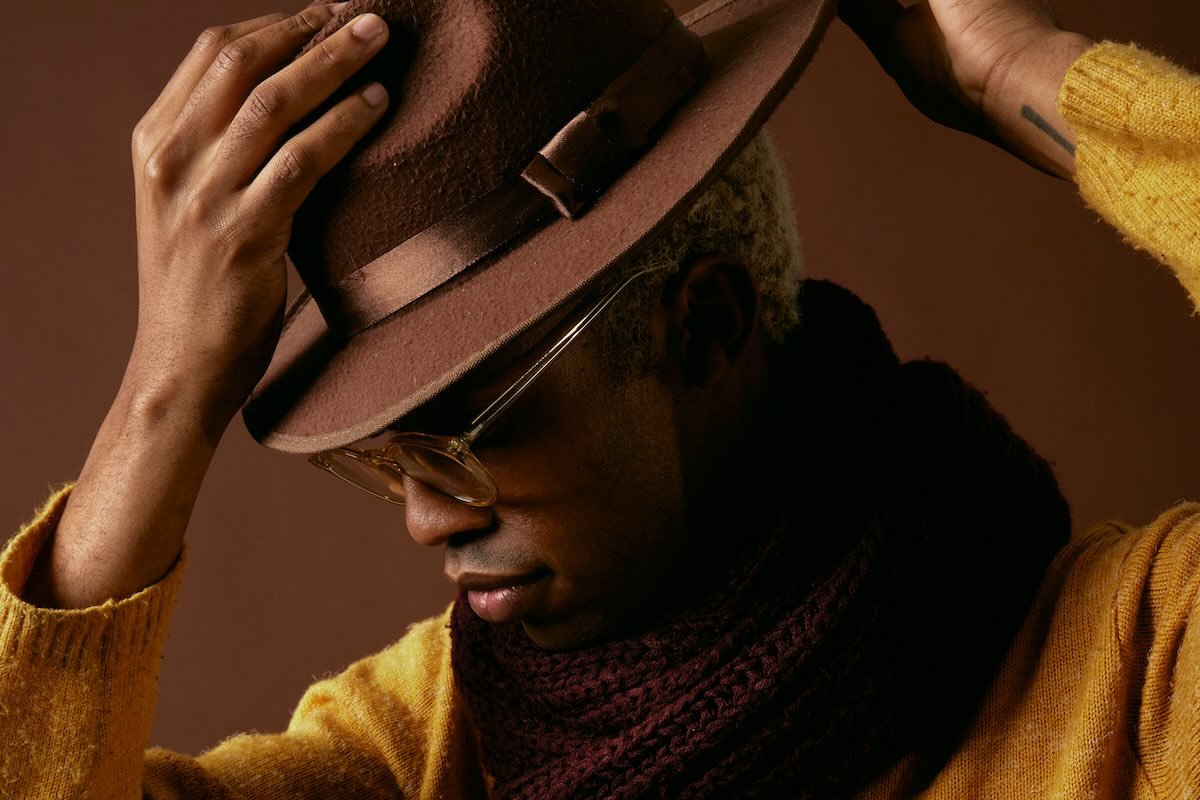 Person in clear-framed glasses, a mustard sweater and burgundy knit scarf, tipping a brown felt fedora over short bleached-blonde hair against a brown backdrop