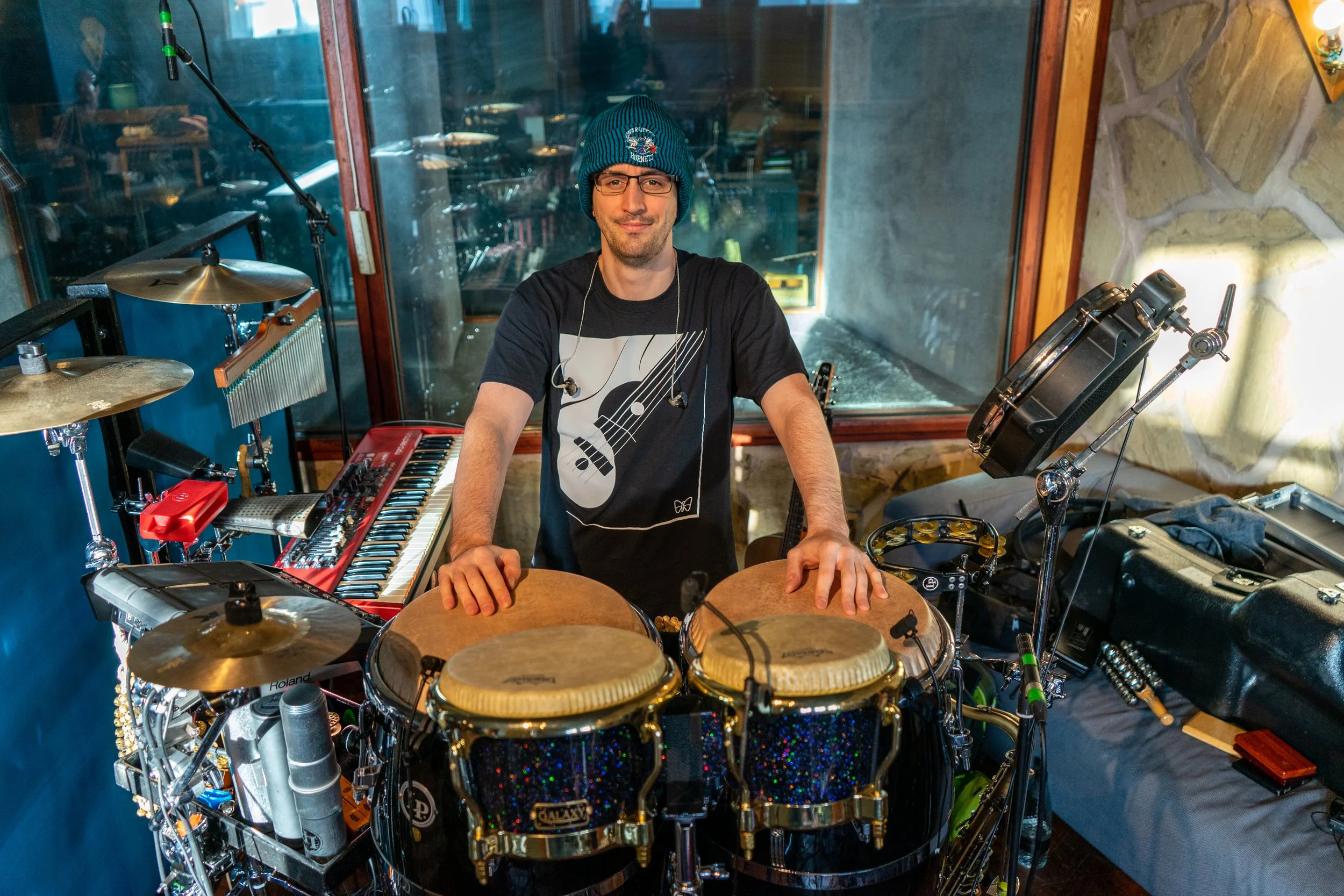A man in a beanie and glasses standing behind a set of percussion instruments in a recording studio, with a keyboard and other musical equipment around him.