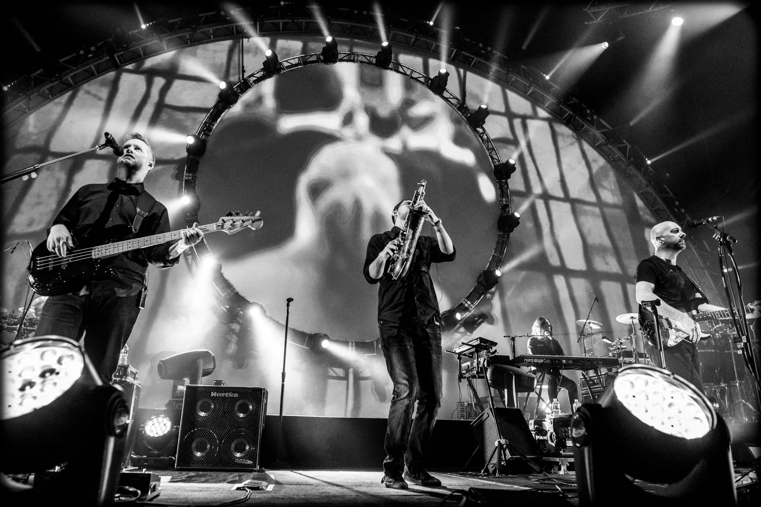 Black and white photo of a band performing on stage, with three musicians playing instruments and a large circular screen behind them displaying abstract visuals.