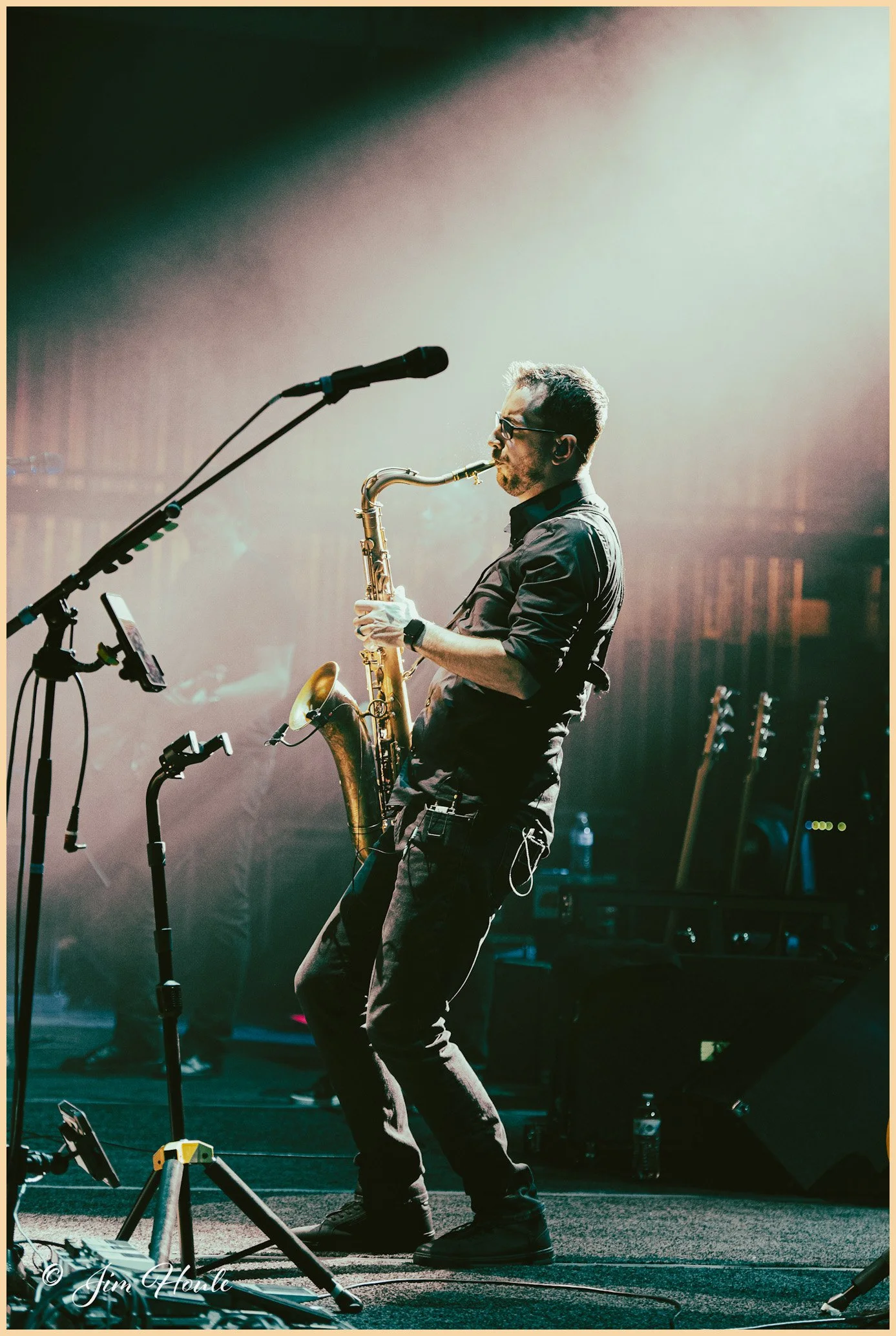 Musician playing saxophone on stage during concert, with microphone and music equipment around him.
