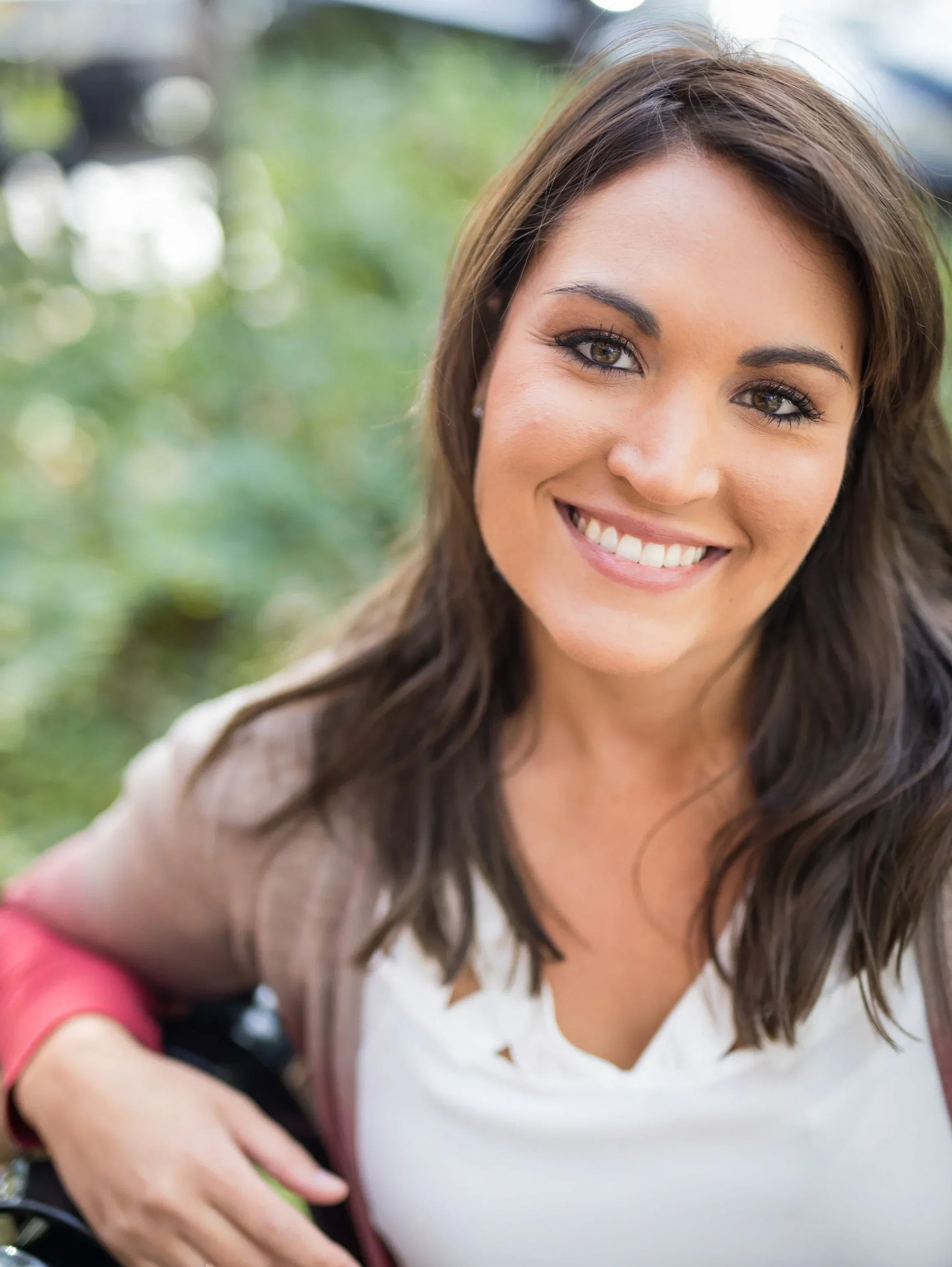 A close-up of a smiling woman with brown hair and blue eyes outdoors with greenery in the background.
