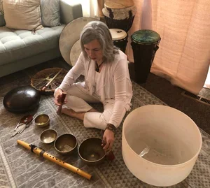 Sherry Strand, Usui Reiki master and holistic health practitioner at Resourced & Resilient, sitting on the floor practicing sound therapy with singing bowl with a flute and drum nearby, in a cozy holistic healing space.
