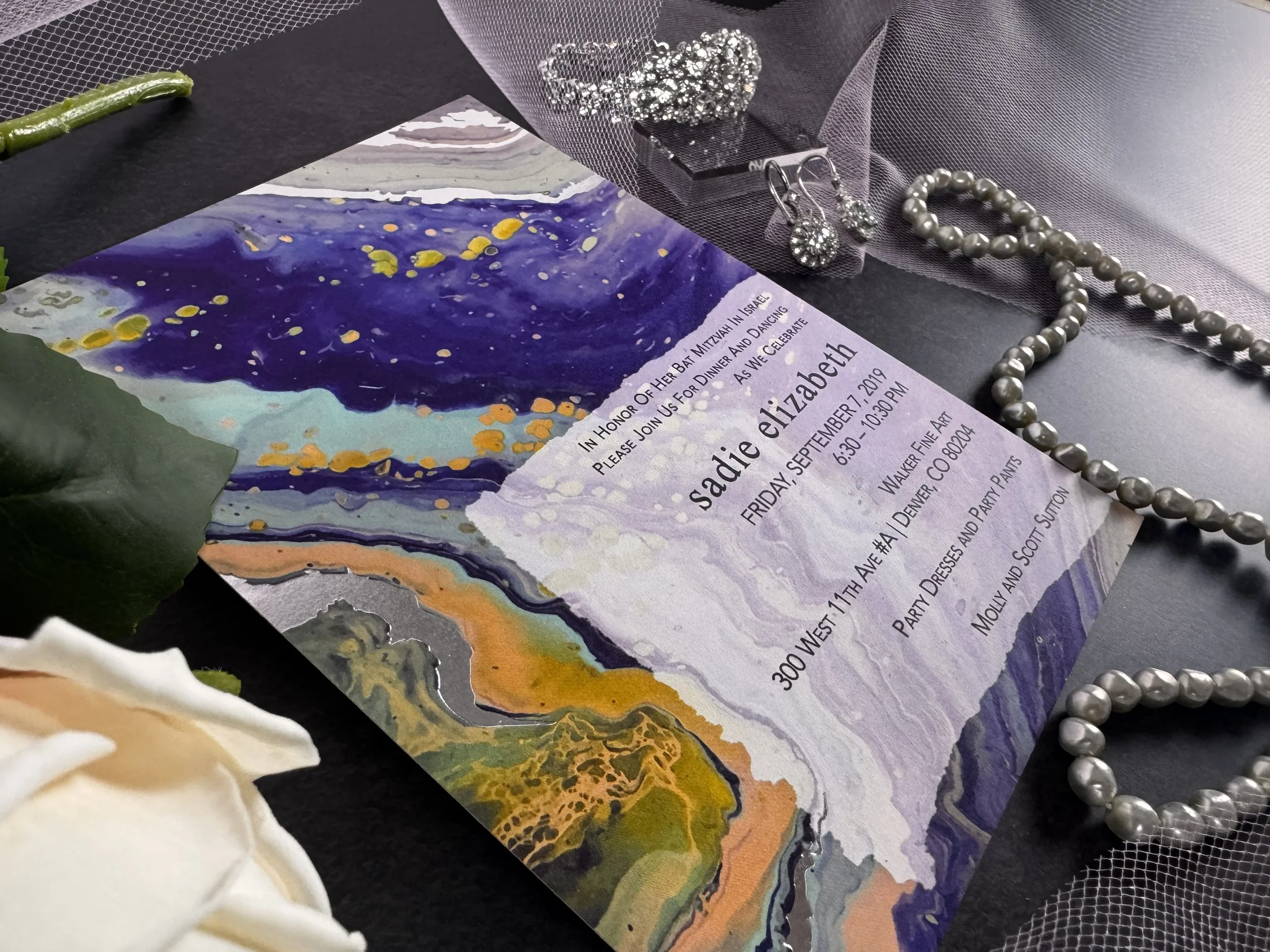 A Bat Mitzvah invitation for Sadie Elizabeth with a colorful abstract background, decorated with pearl jewelry, rhinestone earrings, and a mesh fabric overlay.