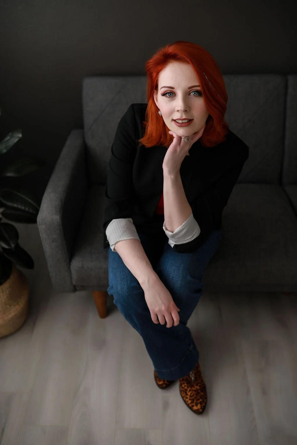 A woman with red hair sitting on a dark gray couch, near a potted plant, resting her chin on her hand, wearing a black jacket, jeans, and leopard print shoes.