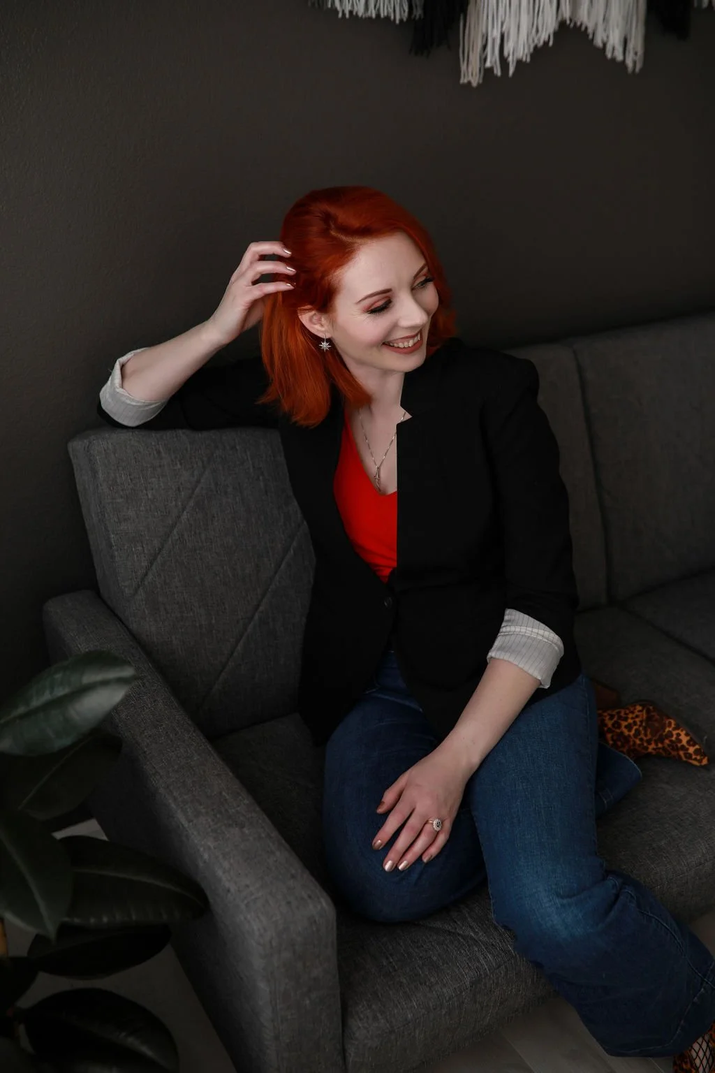 A woman with red hair sitting on a gray sofa, smiling and touching her hair, wearing a black blazer, red top, and blue jeans, with a plant in the foreground.