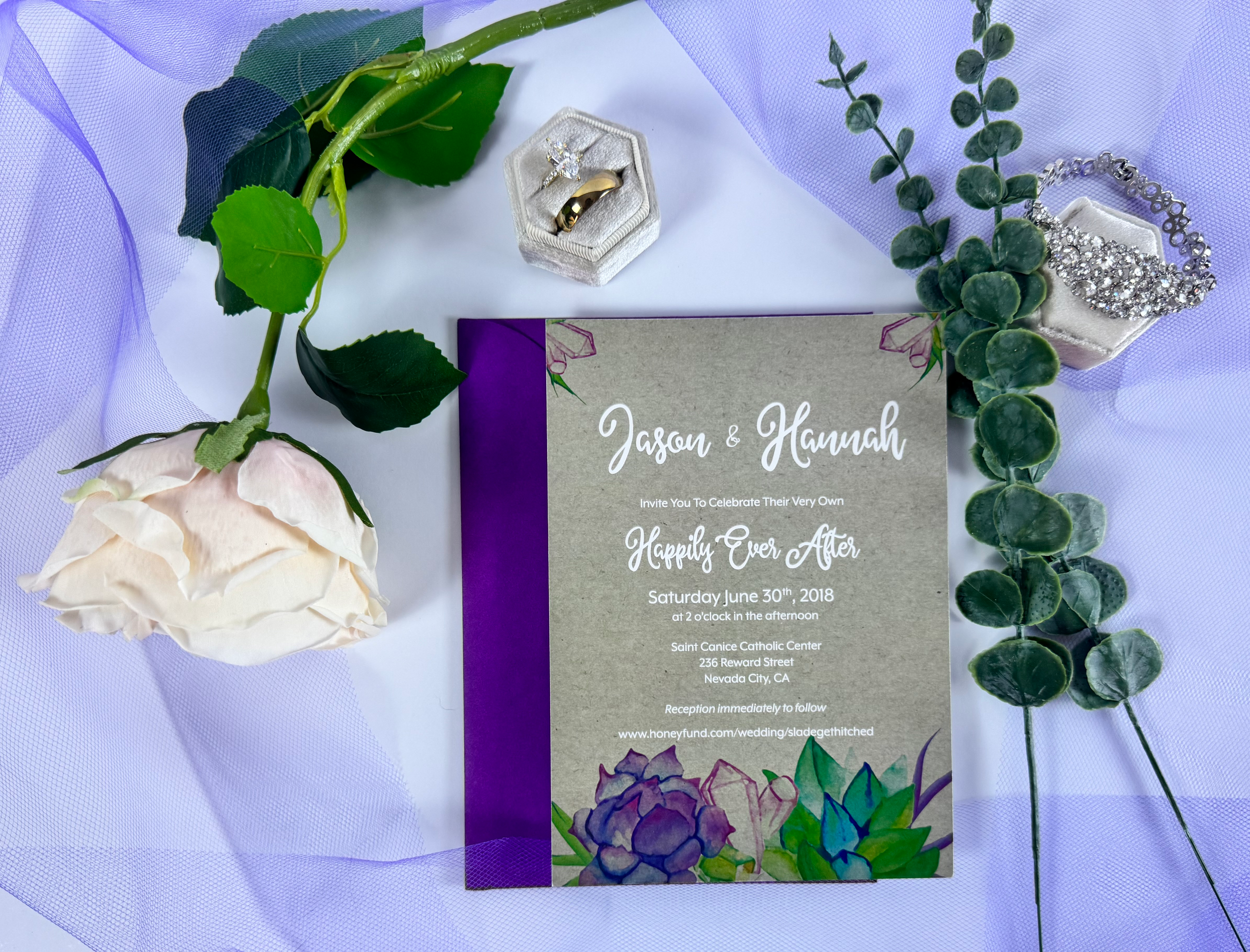 Wedding invitation card surrounded by green leaves, a white rose, and purple flowers. The card has purple and gray colors, and details about the wedding of Jason and Hannah on June 30, 2018, at Saint Canice Catholic Center in Nevada City, CA. There are also jewelry items including a diamond ring and a diamond bracelet.