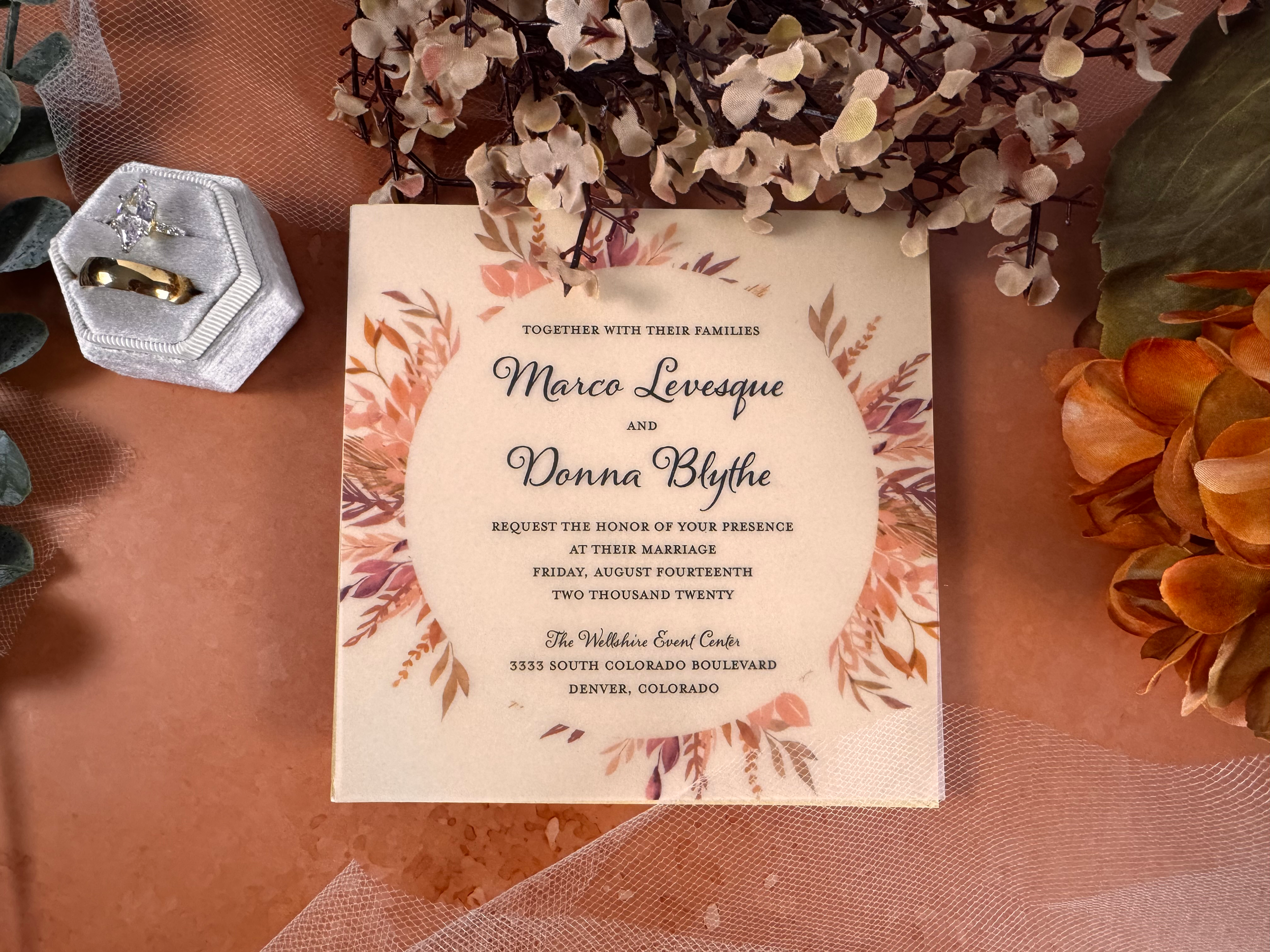 Wedding invitation card with floral design and text details, surrounded by decorative elements including rings in a display box, artificial flowers, and mesh fabric on a pink background.