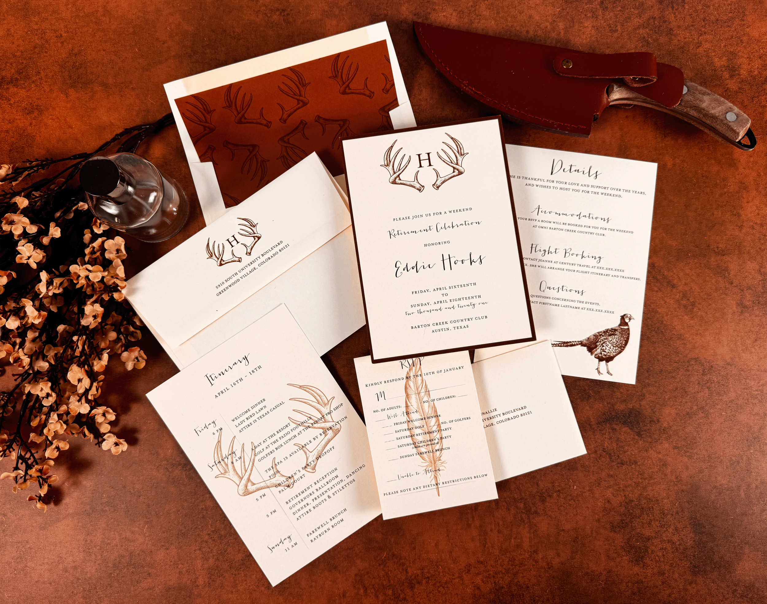 Invitation cards with a deer antler design and event details, a brown leather knife sheath, a clear glass bottle, and orange blossoms on a reddish-brown surface.