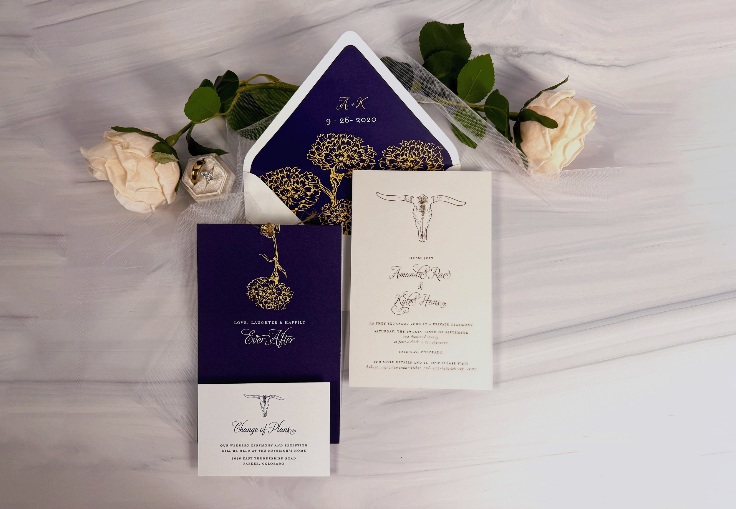 Wedding invitation suite with dark blue and white cards, gold floral designs, and a white rose, arranged on a white surface with tulle and green leaves.