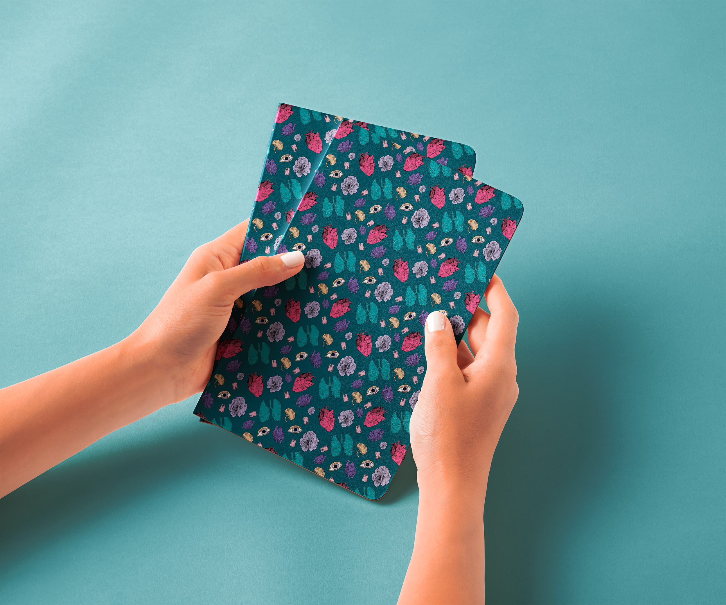 Person holding two notebooks with dark teal covers decorated with colorful floral and eye patterns, against a light teal background.