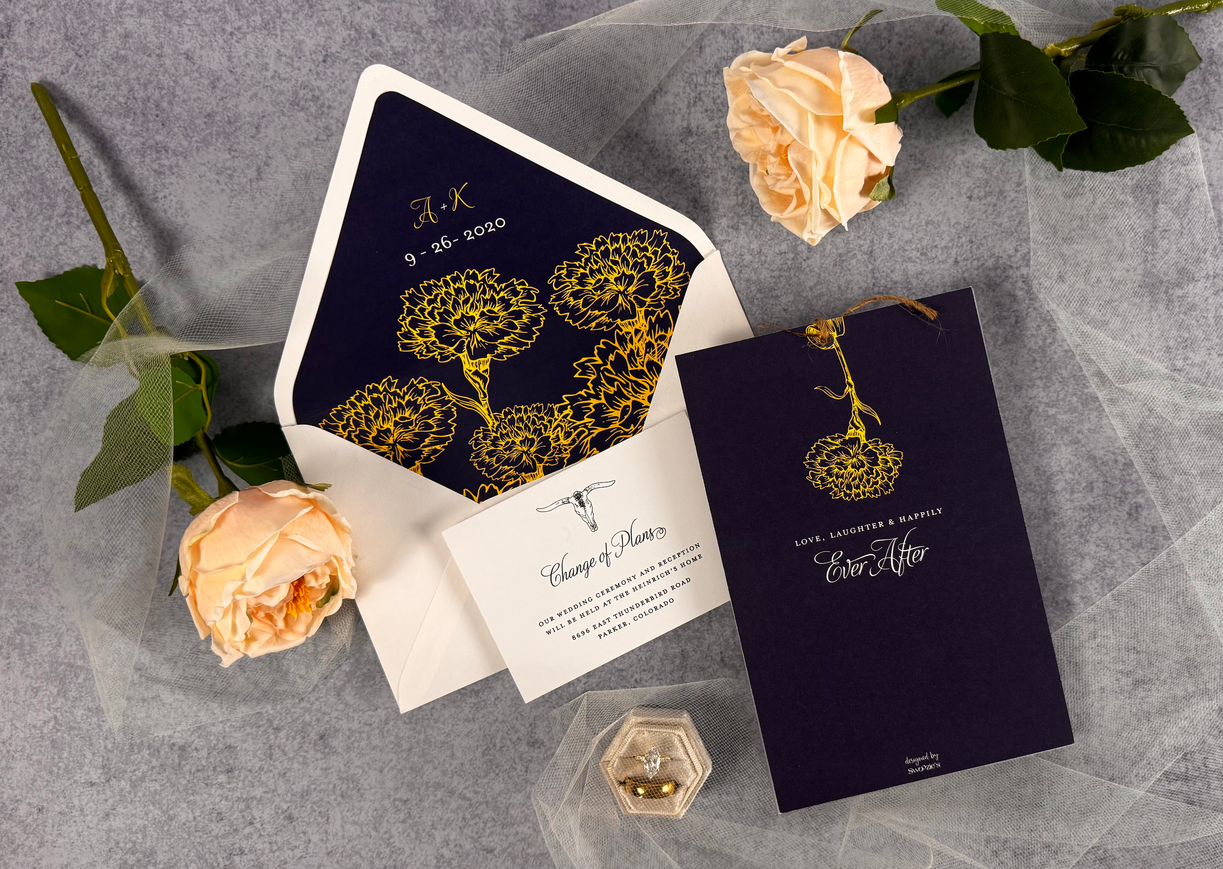 Wedding invitation suite with dark navy and gold floral designs, accompanied by light peach roses and a hexagonal gold ring box on a gray surface with sheer fabric