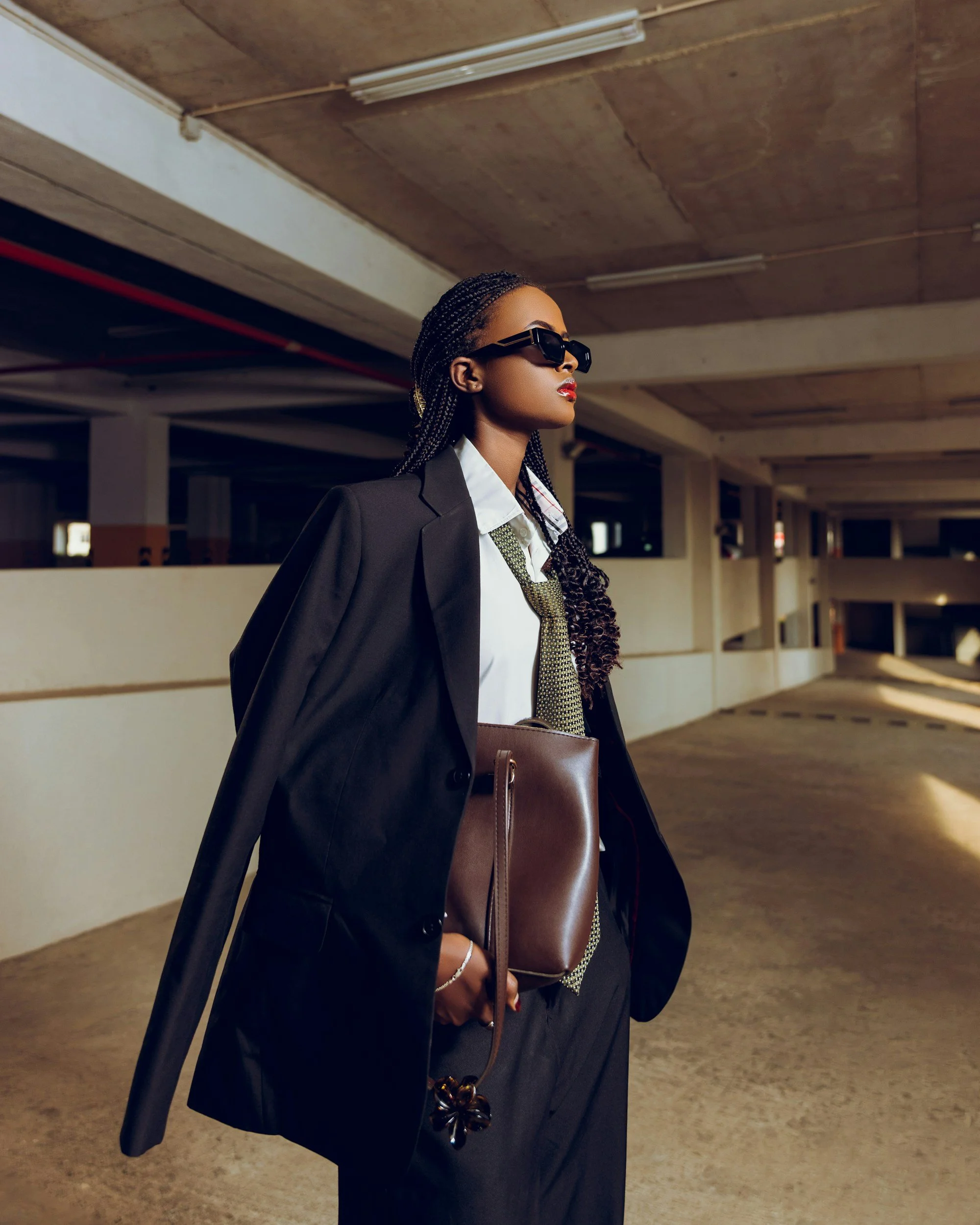 A stylish woman with braided hair wearing sunglasses, a black blazer draped over her shoulders, a white shirt, and a patterned tie, holding a brown handbag, walking through a parking garage.