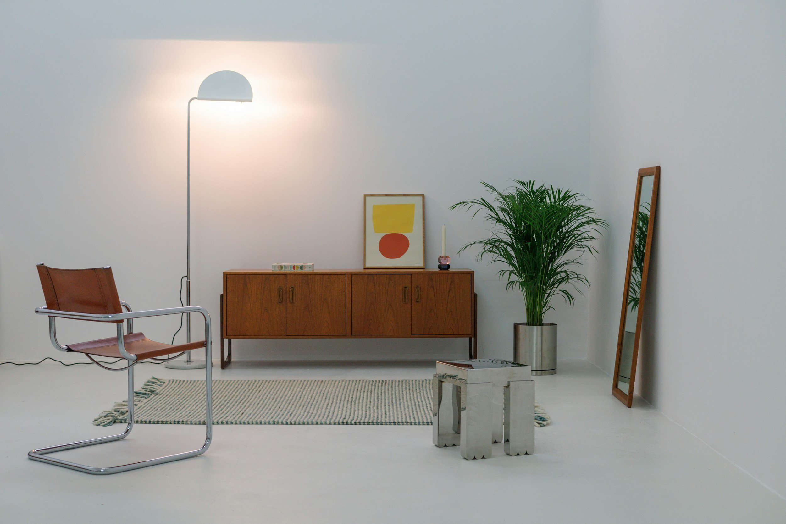 Minimalist interior with a brown chair, floor lamp, wooden sideboard, framed artwork, potted plant, mirror, small side tables, and a rug on white walls and floor.