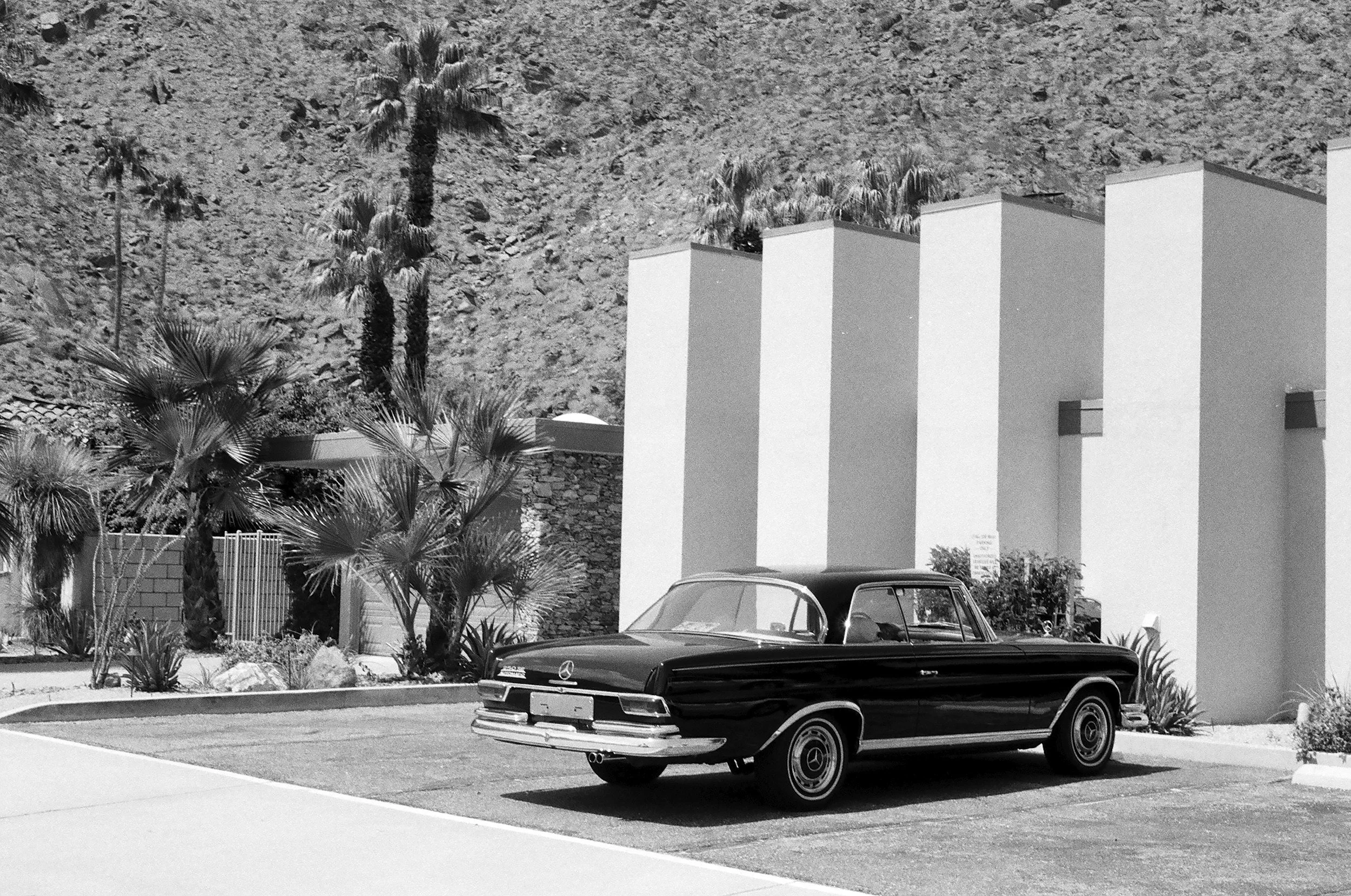 Black vintage Mercedes-Benz car parked on a driveway in front of modern white buildings with flat roofs and palm trees, against a rocky hillside.