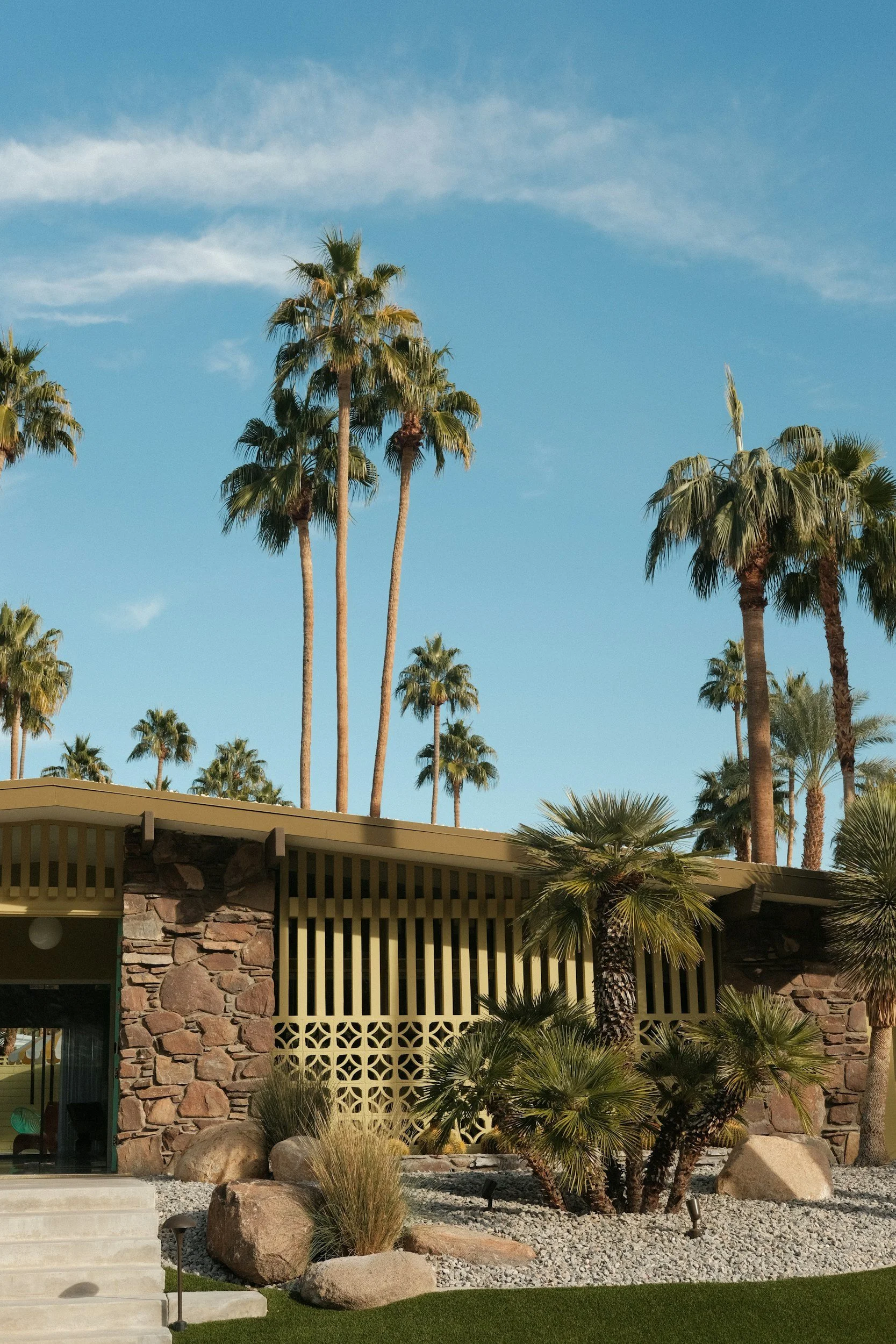 A mid-century modern building with a stone facade, surrounded by desert plants and tall palm trees, under a blue sky with some wispy clouds.