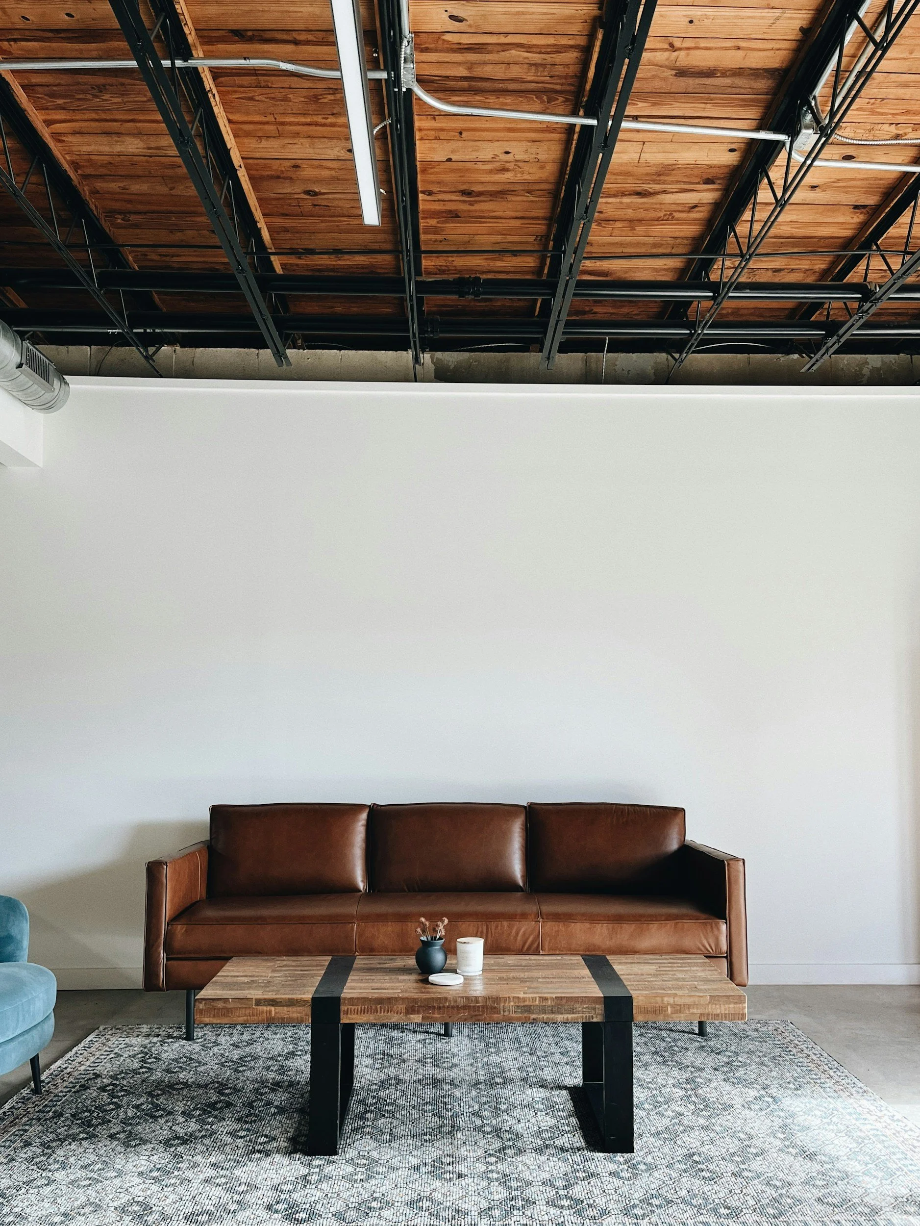 Modern lounge with brown leather sofa, wooden coffee table, blue armchair, patterned rug, and exposed ceiling with wooden beams and industrial pipes.