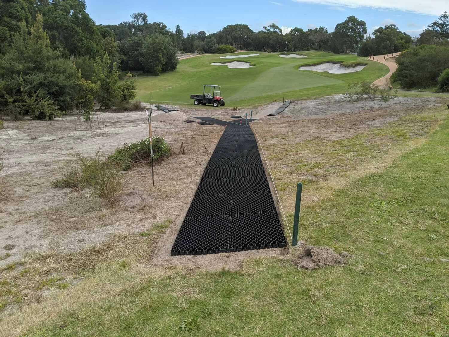 Golf course under construction with a black pathway, construction materials, and a golf cart on the fairway.