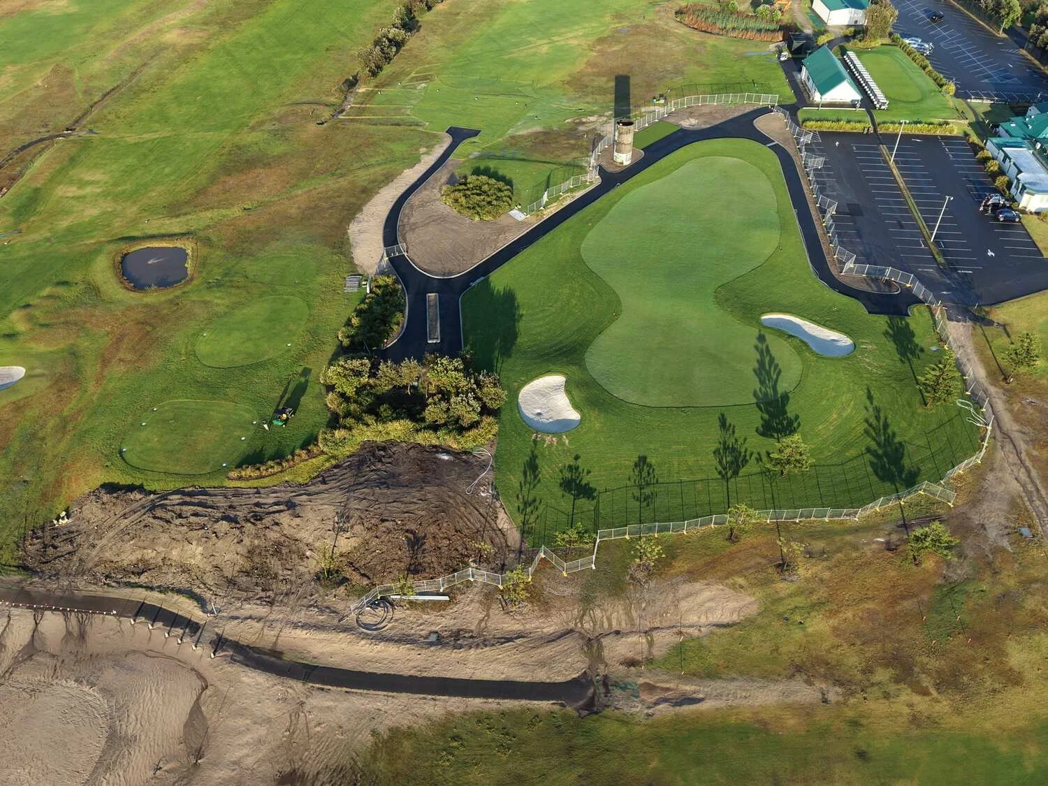 Aerial view of a golf course with multiple sand bunkers, a small pond, trees, and a parking lot with green-roofed facilities.