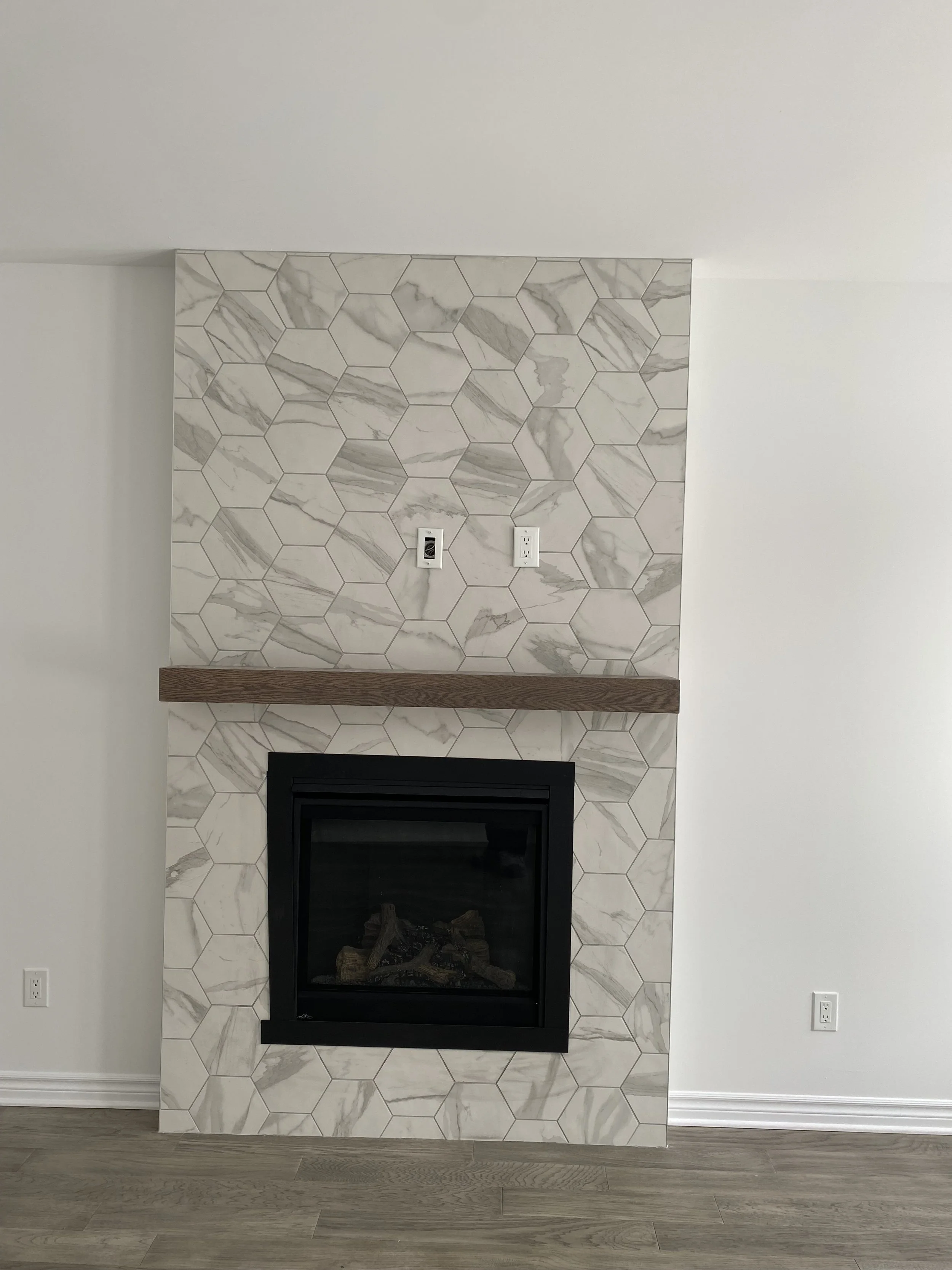 Modern fireplace with a black framed glass front, set in a wall surrounded by large white marble tiles with gray veining, and a wooden mantel shelf above, situated in a minimalistic room with light hardwood flooring and white walls.