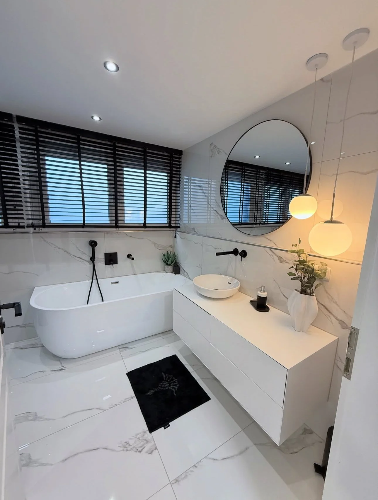 Modern bathroom with white marble walls and floor, black window blinds, a white bathtub, a white vanity with a vessel sink, round mirror, black fixtures, potted plants, and pendant lights with warm glow.