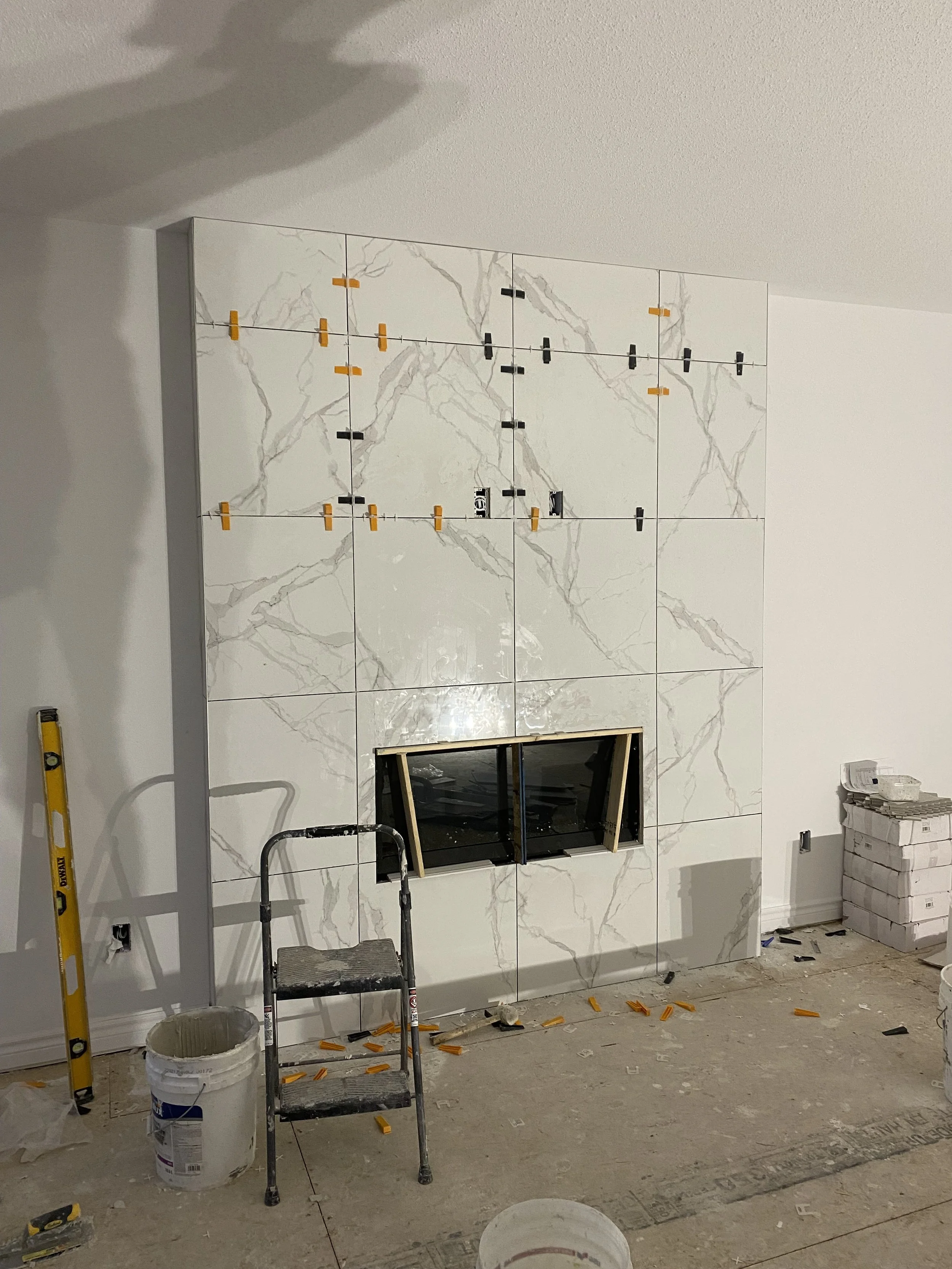 Interior wall under construction with large marble or marble-look tiles, small black and yellow spacers, and a built-in fireplace opening, with construction tools and materials around.