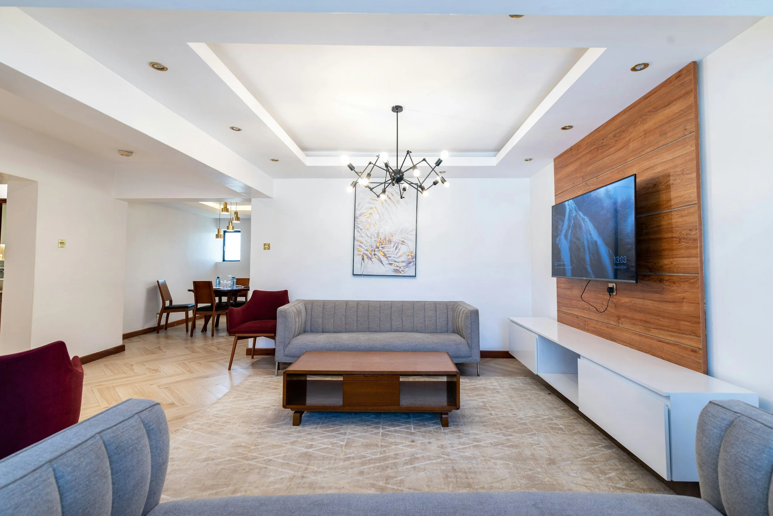 Modern living room with a grey sofa, wooden coffee table, wall-mounted flat-screen TV on a wood-paneled accent wall, and artwork above the sofa. Adjacent dining area with a wooden table and chairs, and a contemporary chandelier hanging from the ceiling.