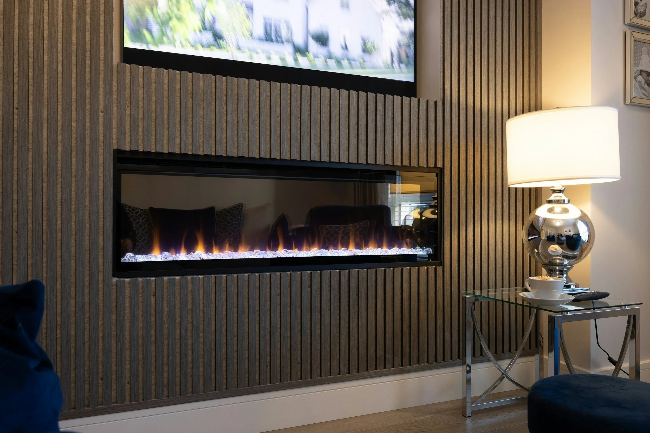 A living room with a modern wooden slat wall, a horizontal electric fireplace, a table with a large silver lamp, a cup, and a remote control, and a flat-screen TV mounted above the fireplace.