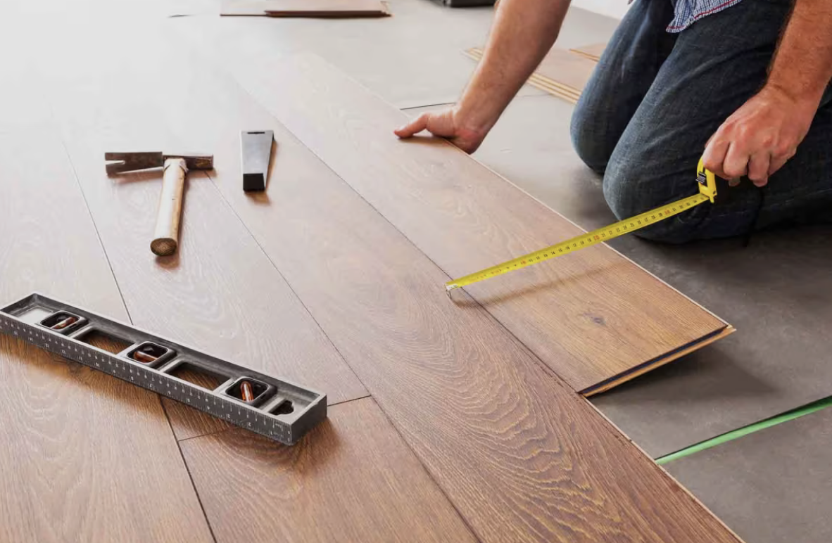 Person kneeling on the floor measuring a wooden plank with a yellow tape measure, with tools like a hammer, a two-in-one square, and a level nearby.