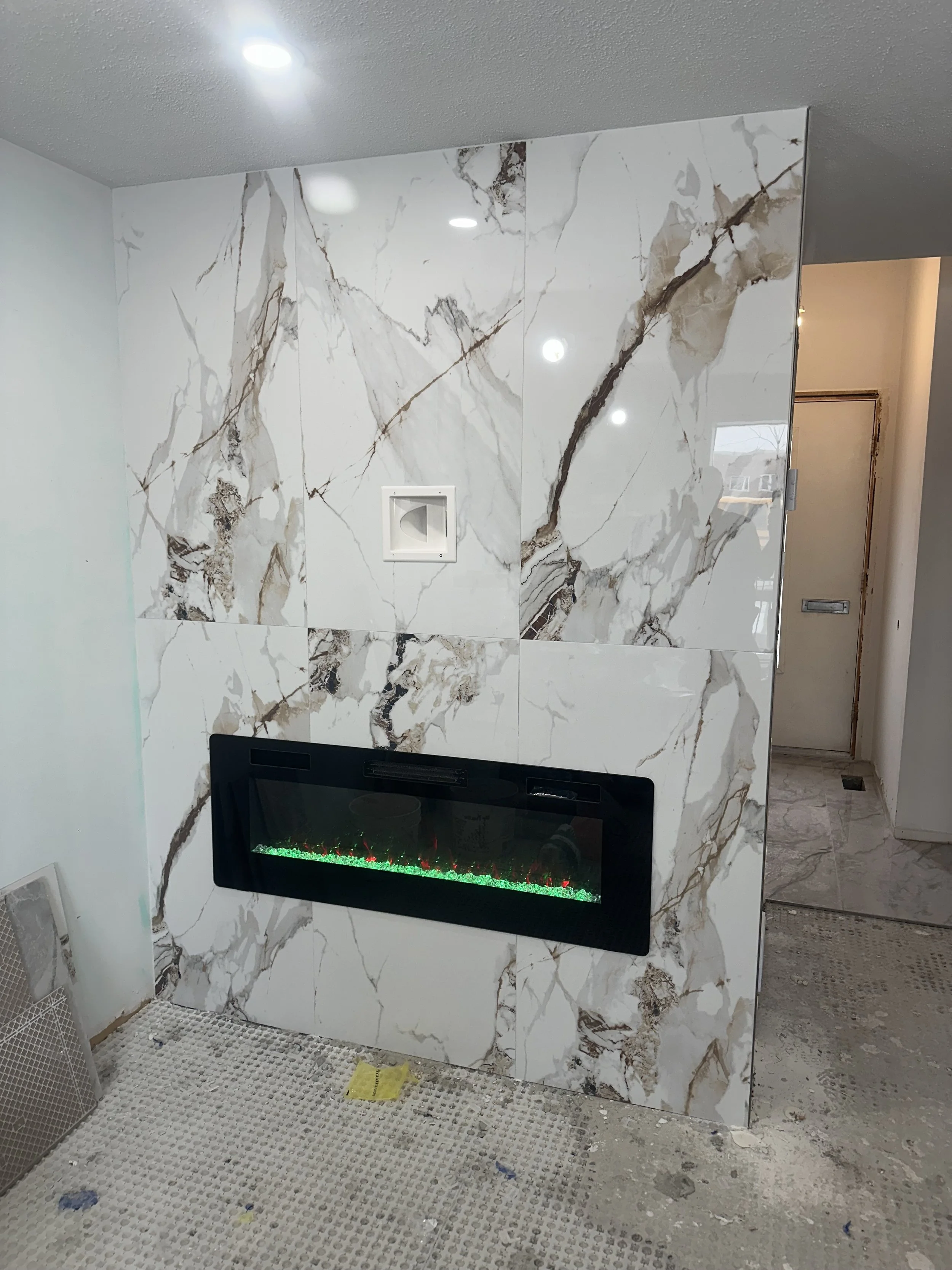 Interior view of a wall with large white marble tiles accented with brown and gray veining. An electric fireplace with a black frame and green and red flames is built into the wall. The ceiling has recessed lighting, and the adjacent room has a door 