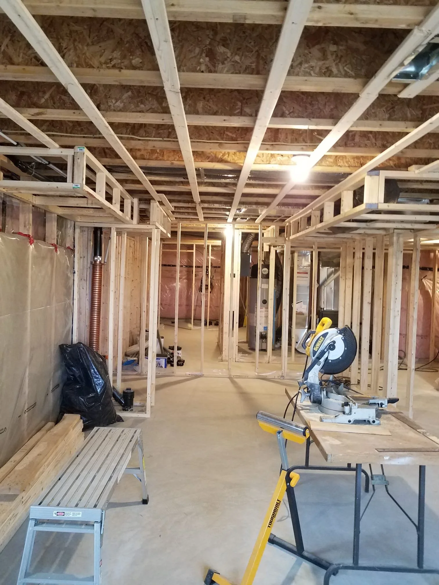 Interior of a house under construction with exposed wooden framing, a work table with a miter saw, a yellow portable work stand, a black plastic bag, and a bench, with lighting illuminating the area.