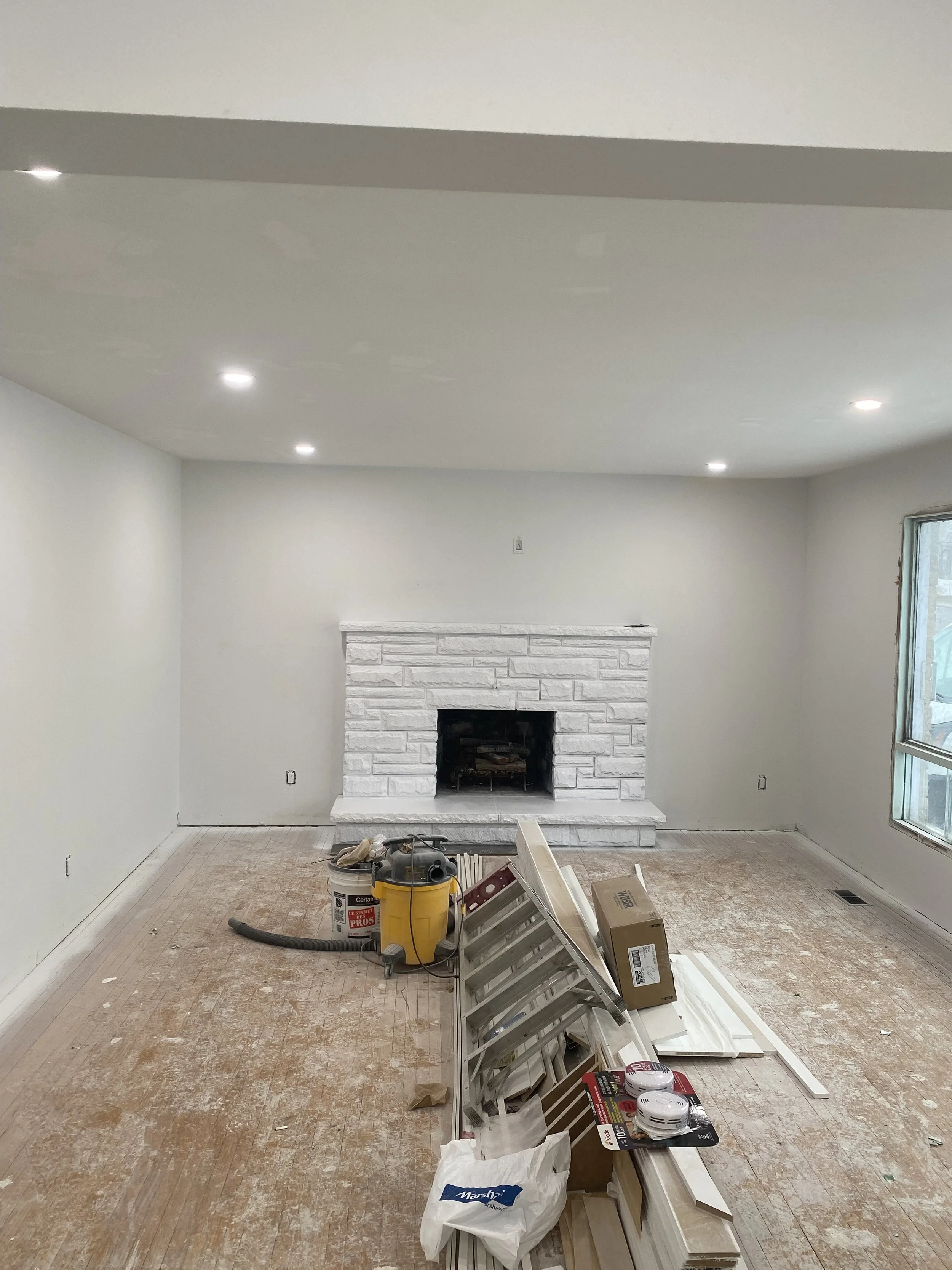 Room under renovation with a white brick fireplace, a window on the right, and construction supplies on the floor including a ladder, a yellow vacuum, boxes, and other tools.