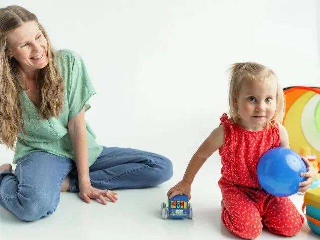 Children and adults playing with a large transparent ball decorated with colorful dots in a room with a plain wall background.