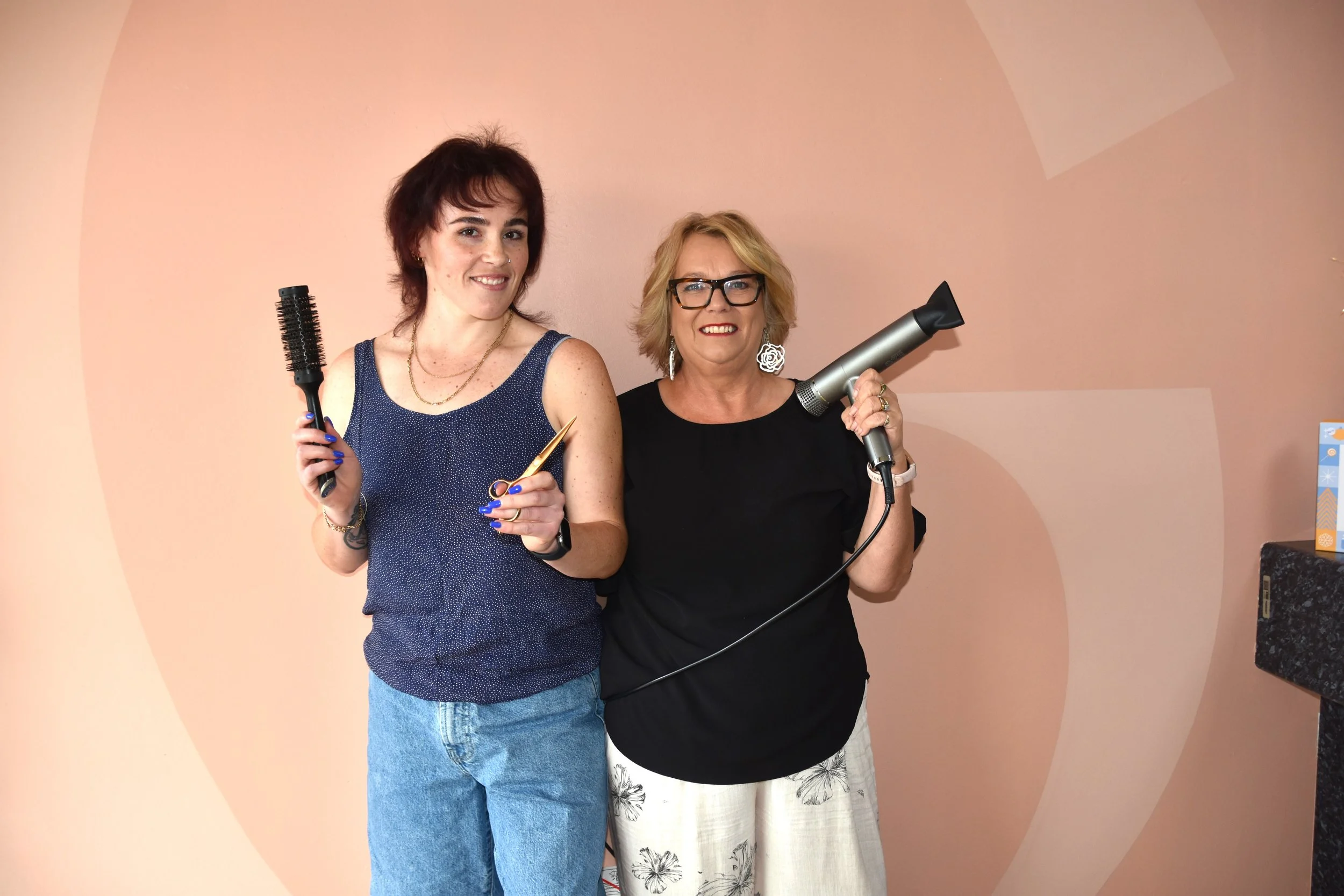 Two women stand together against a pink wall, holding hair styling tools: one with a round brush and scissors, the other with a hairdryer.