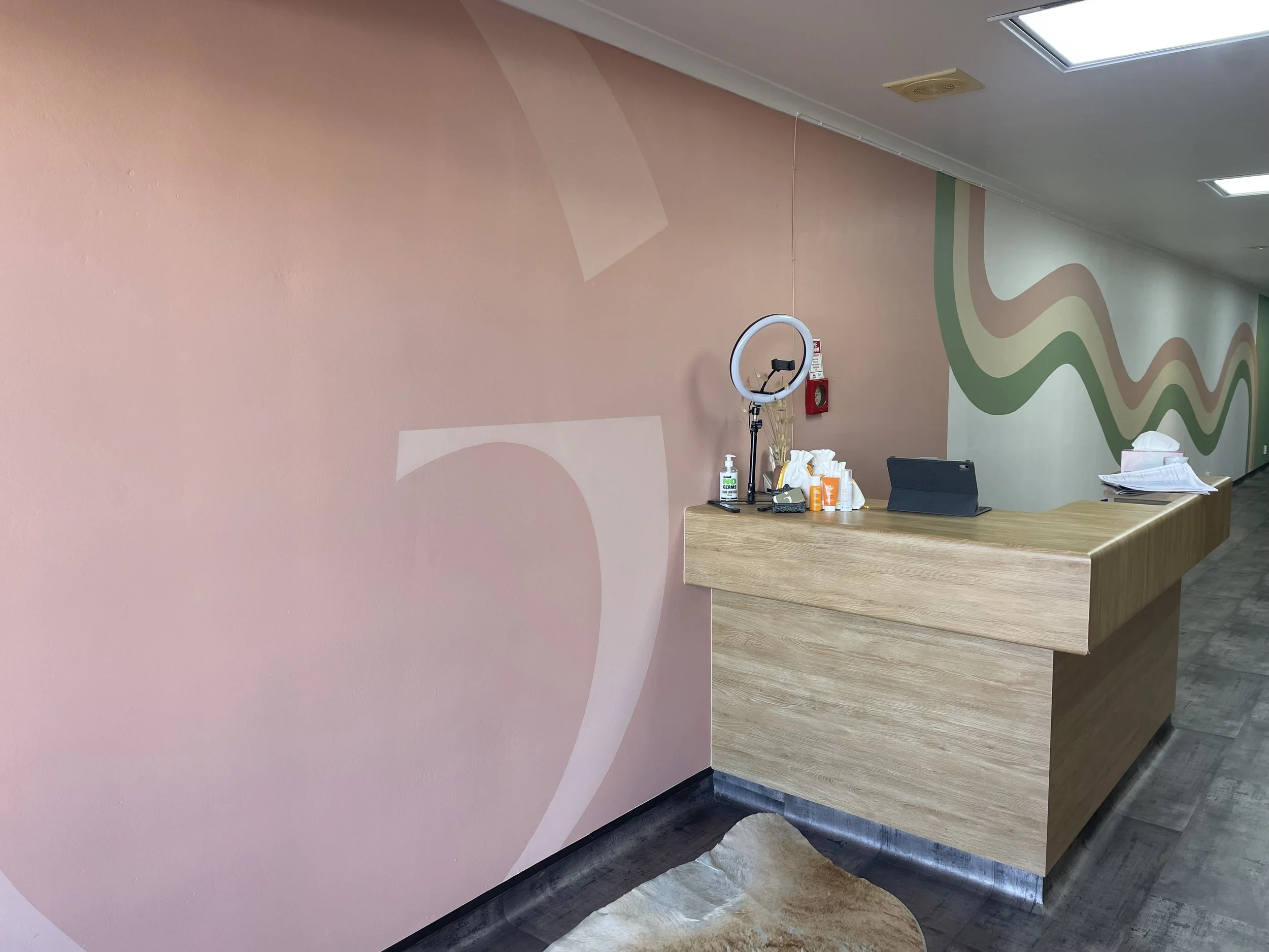 A reception area with a pink and green abstract wall design, a wooden counter with various items including a ring light, sanitizer, tissues, and a tablet, and overhead fluorescent ceiling lights.