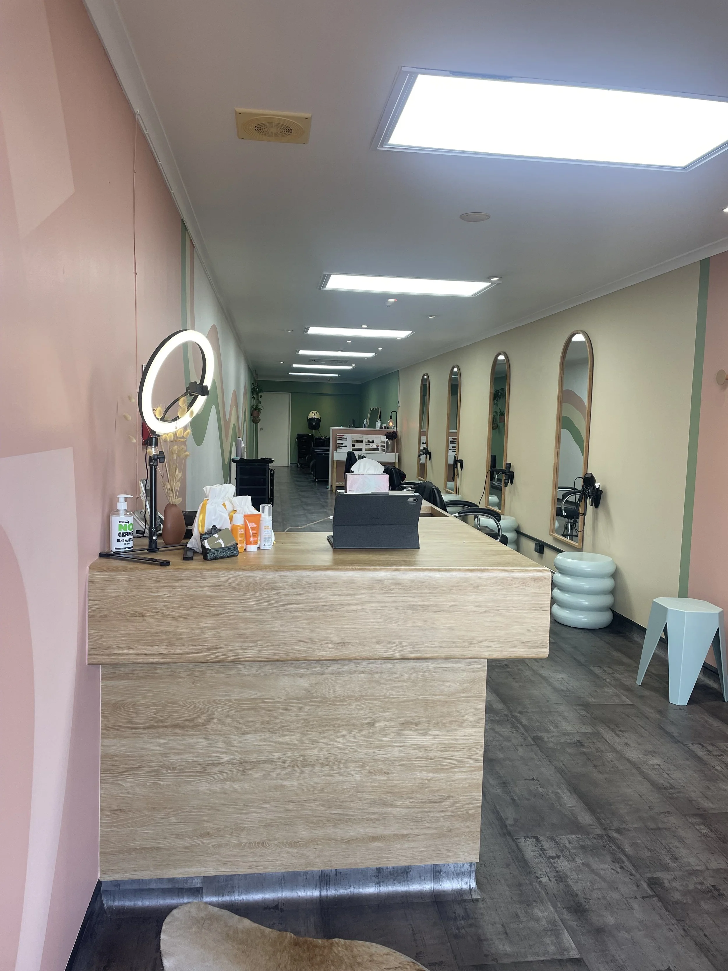 Interior of a hair salon with a light wood reception desk, wall mirrors, and chairs, bright lighting, and pastel-colored walls.