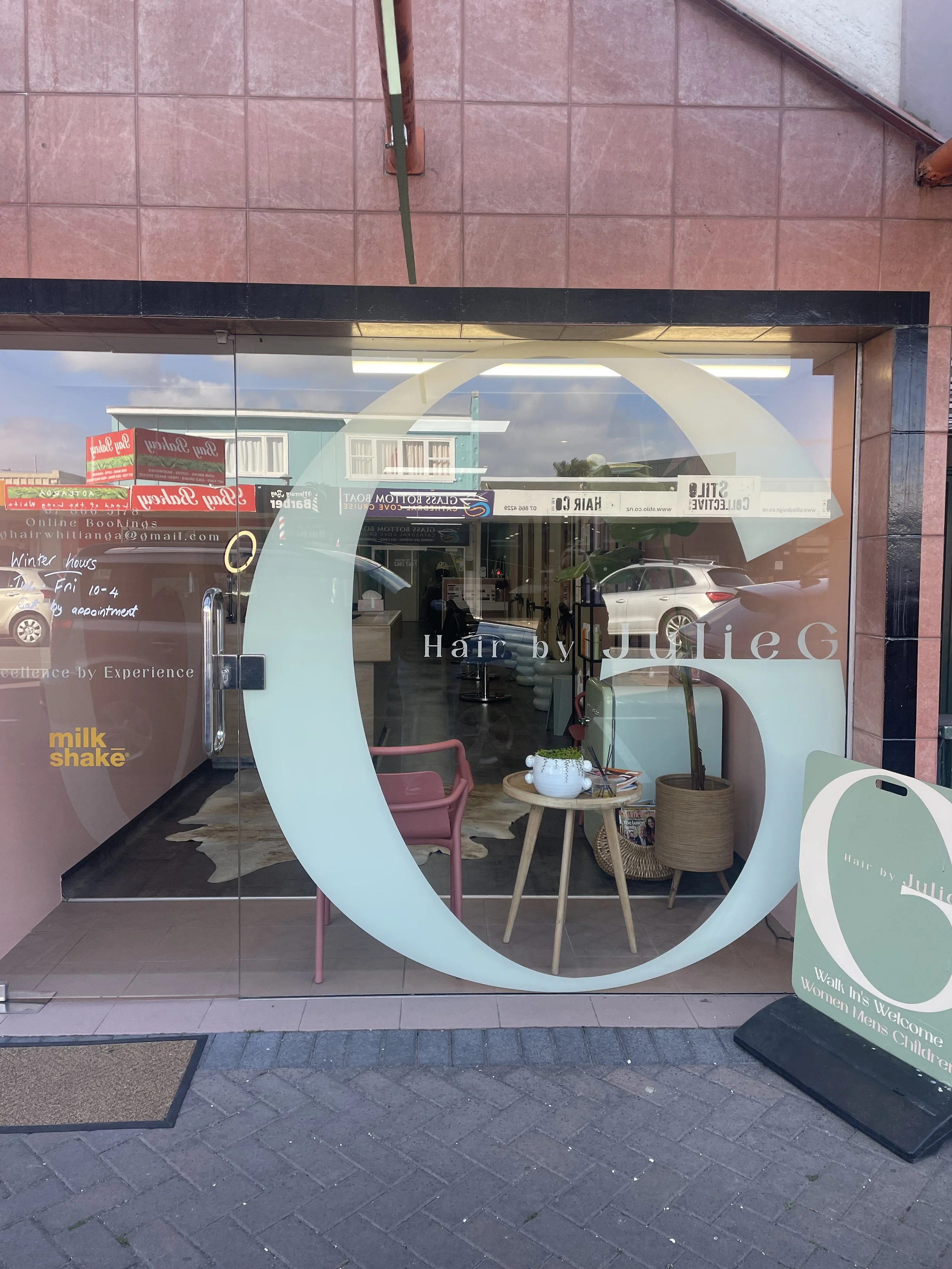 The storefront of a hair salon with large glass windows reflecting the street outside, including parked cars and buildings. Inside, there are white chairs, a small wooden table with a potted plant, and a sign on the door with business hours and conta