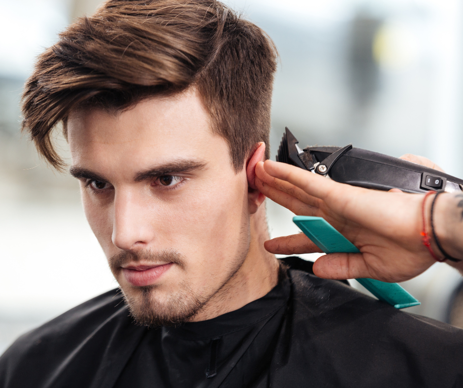 Man getting a haircut at a salon, with hair clippers near his ear, wearing a black cape.