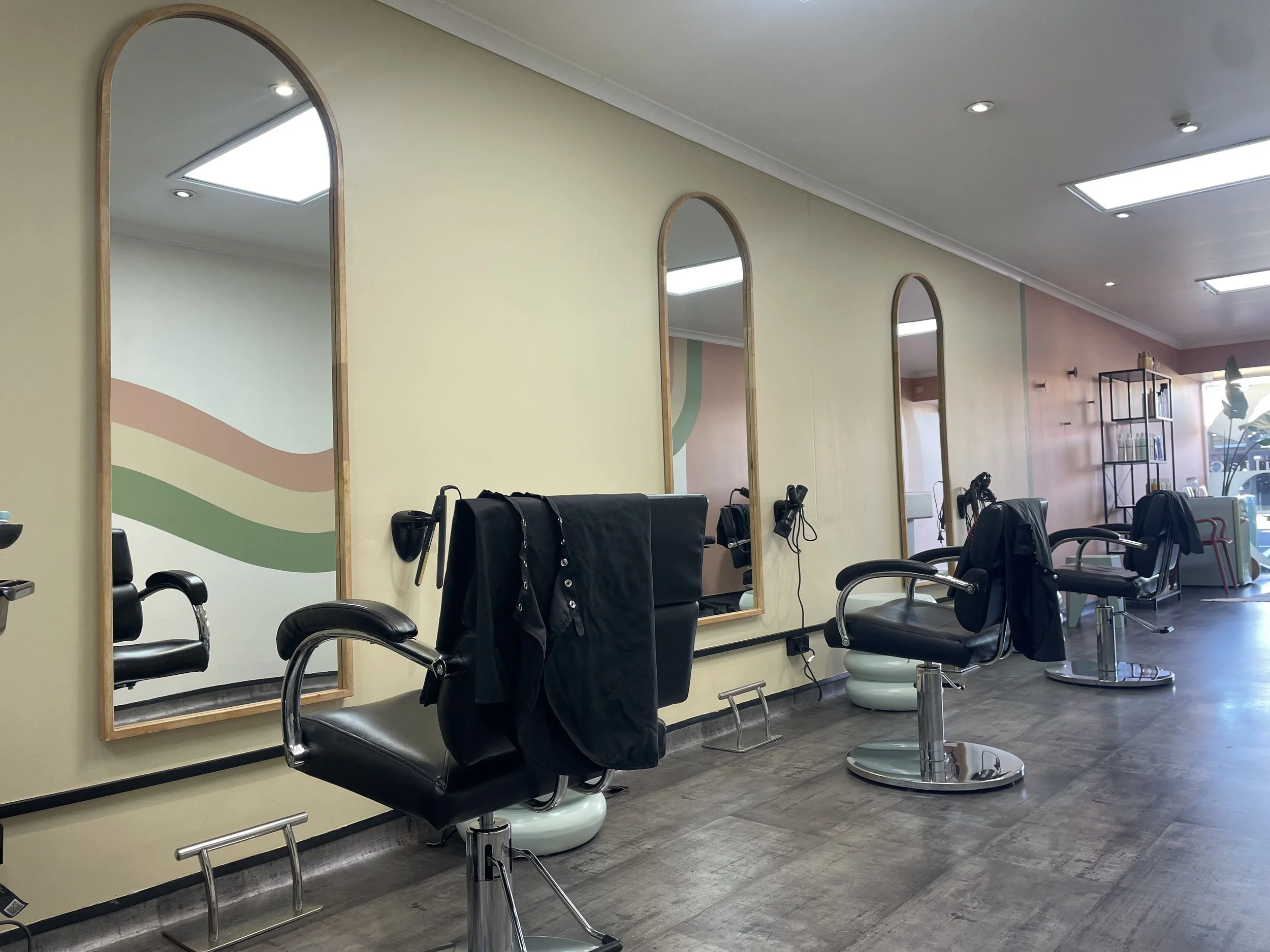 A hair salon with black salon chairs, mirrors, and hair styling tools. The walls are light yellow with decorative mirrors, and there are shelves with products in the background. Large windows let in natural light.