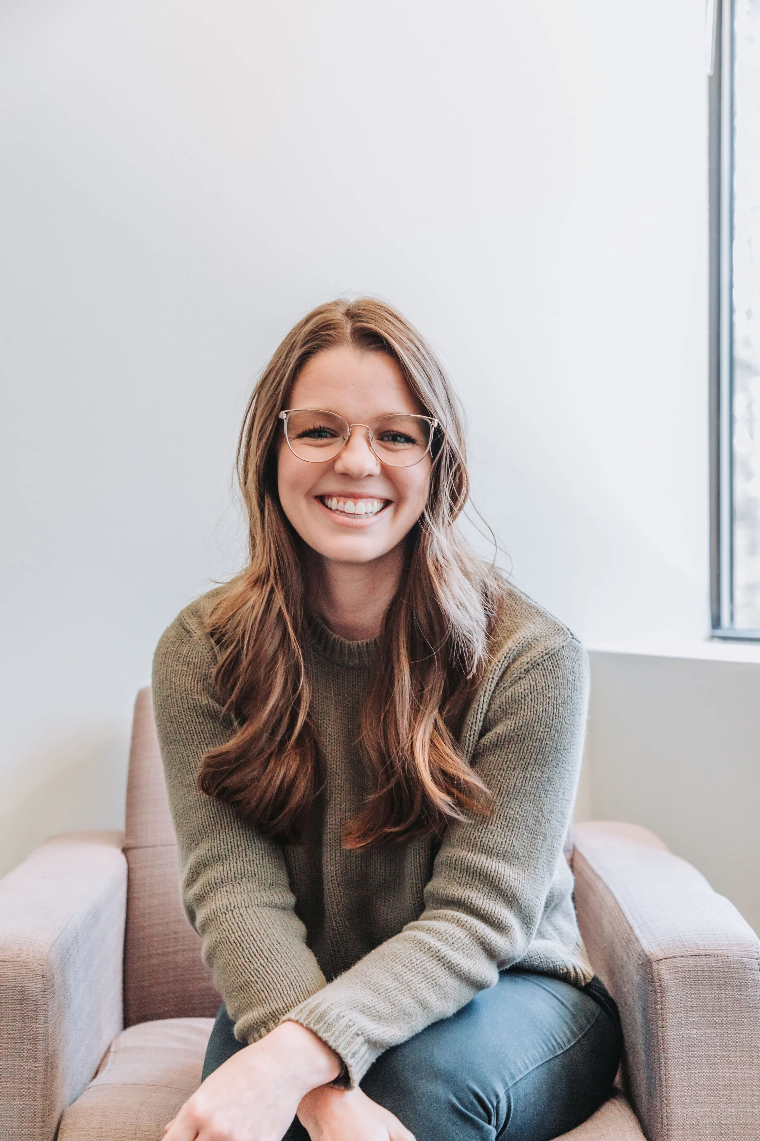 A young woman with long, wavy brown hair and glasses smiling while sitting on a pink armchair near a window.