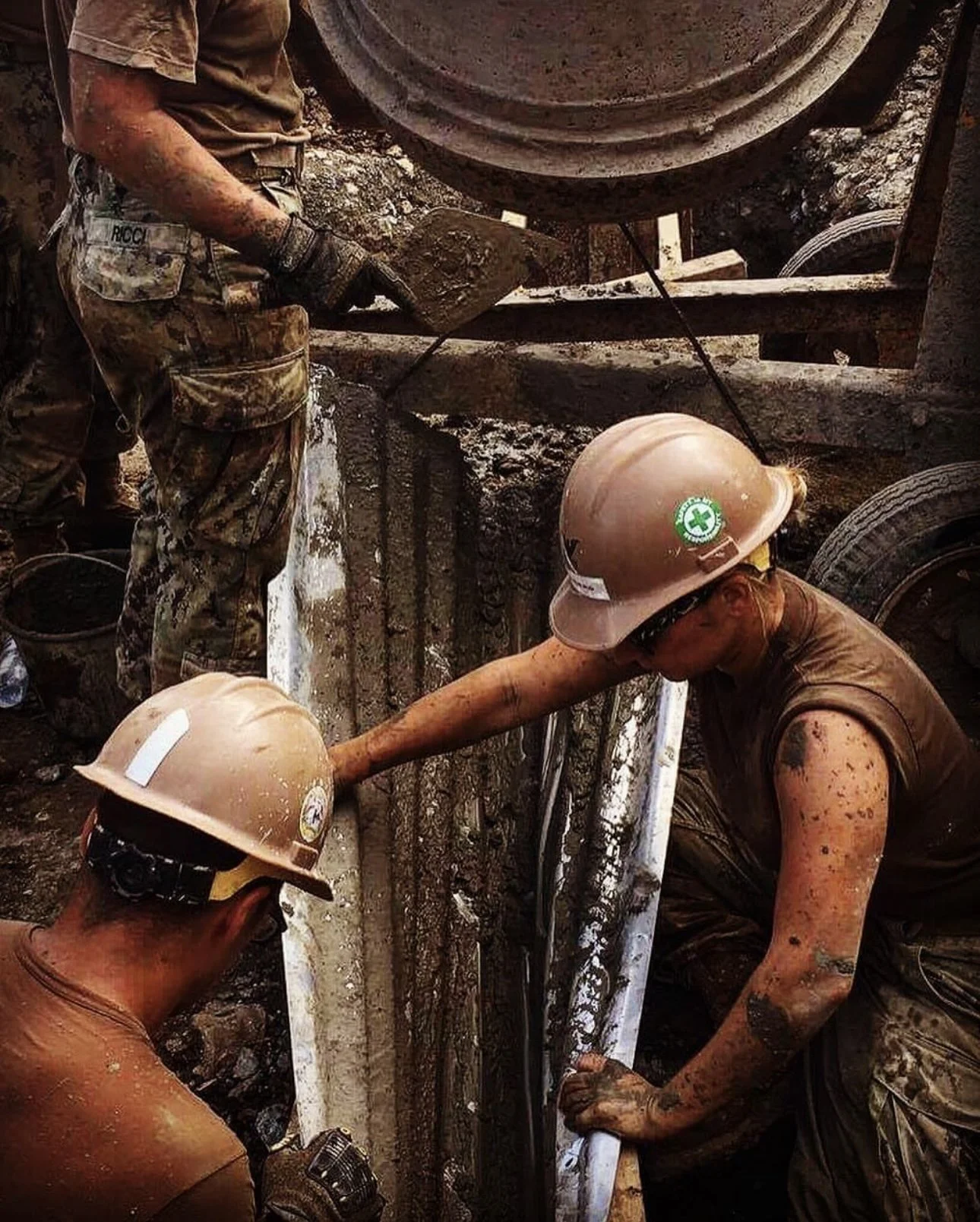 Two workers in safety helmets and muddy clothes working together on a construction site, handling pipes and tools amidst dirt and construction materials.