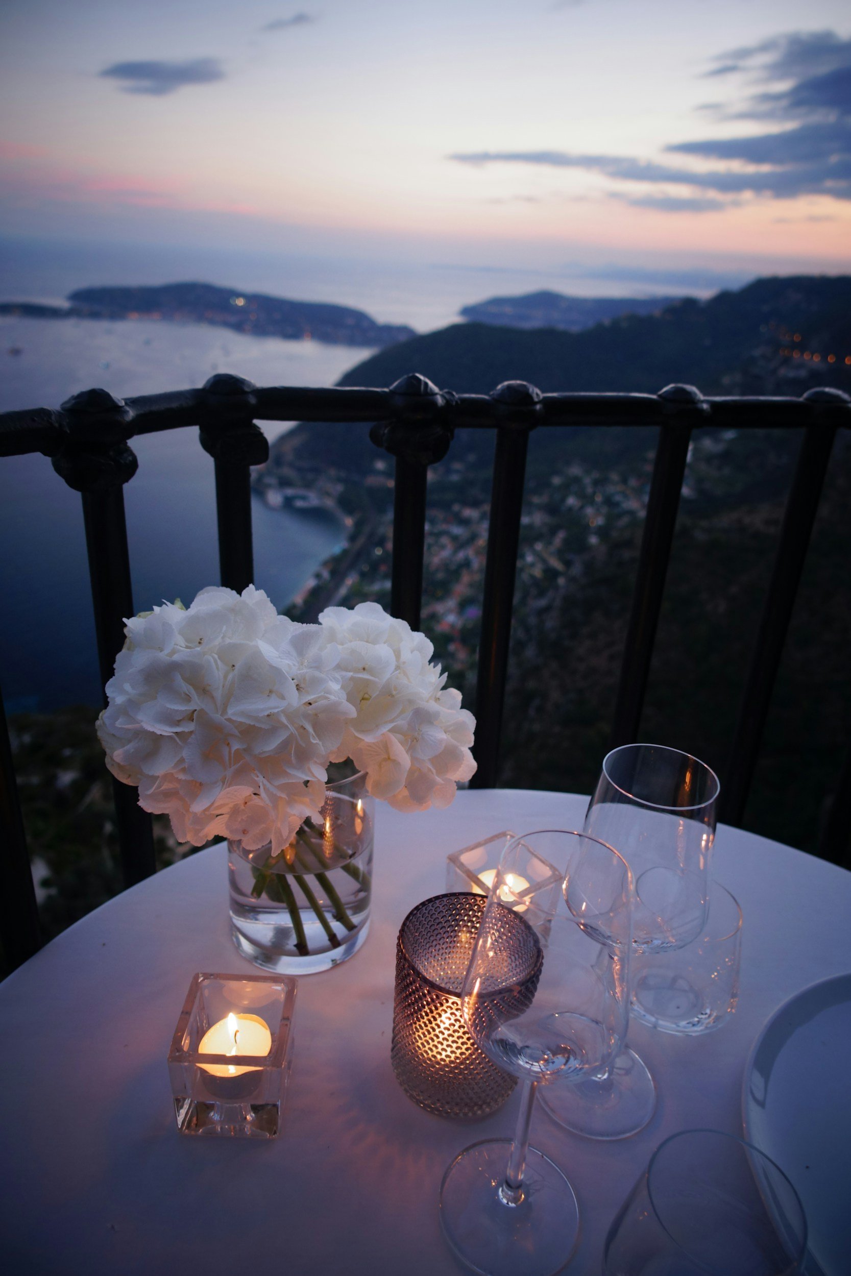 A table set for dinner with a vase of white hydrangeas, lit candles, and empty wine glasses, overlooking a scenic coastal sunset view.