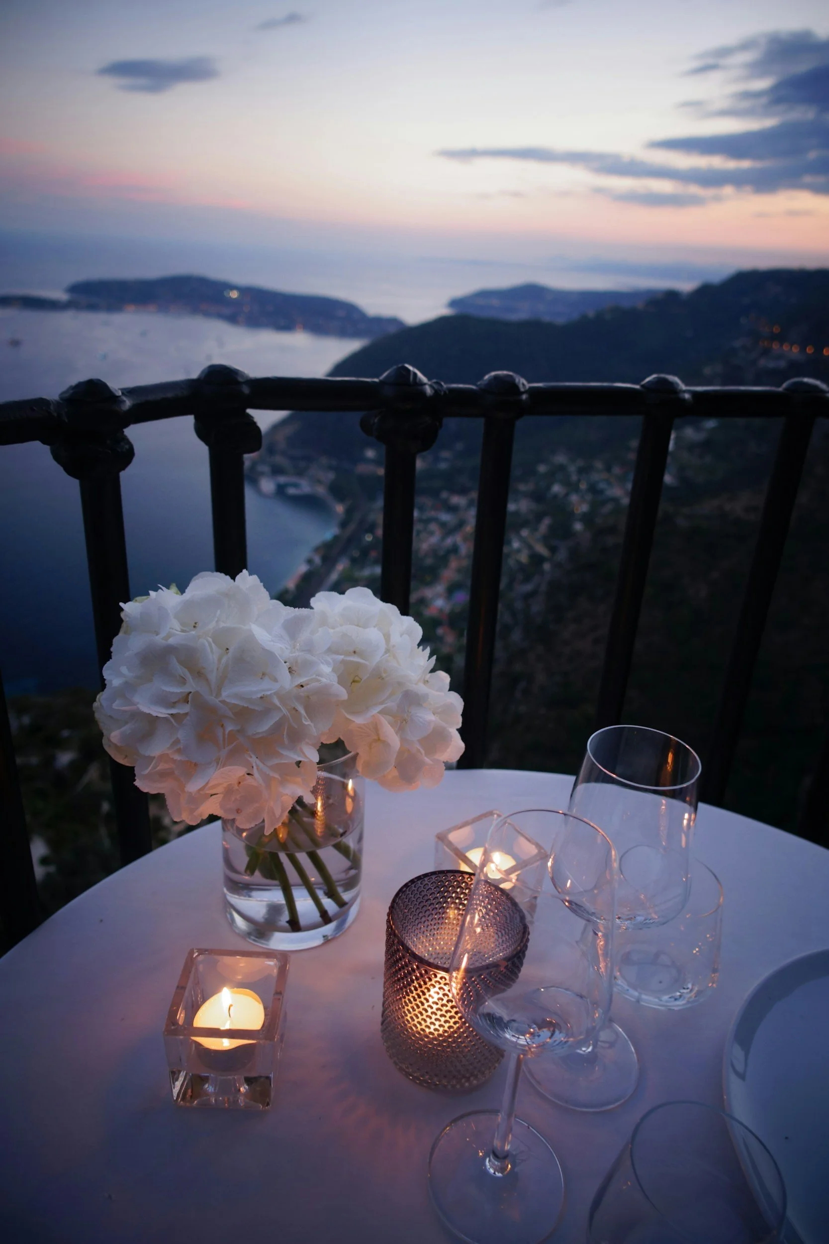 A table set with a vase of white hydrangeas, lit candles, and glassware, overlooking a scenic coastal landscape during sunset, with hills, water, and a distant town.