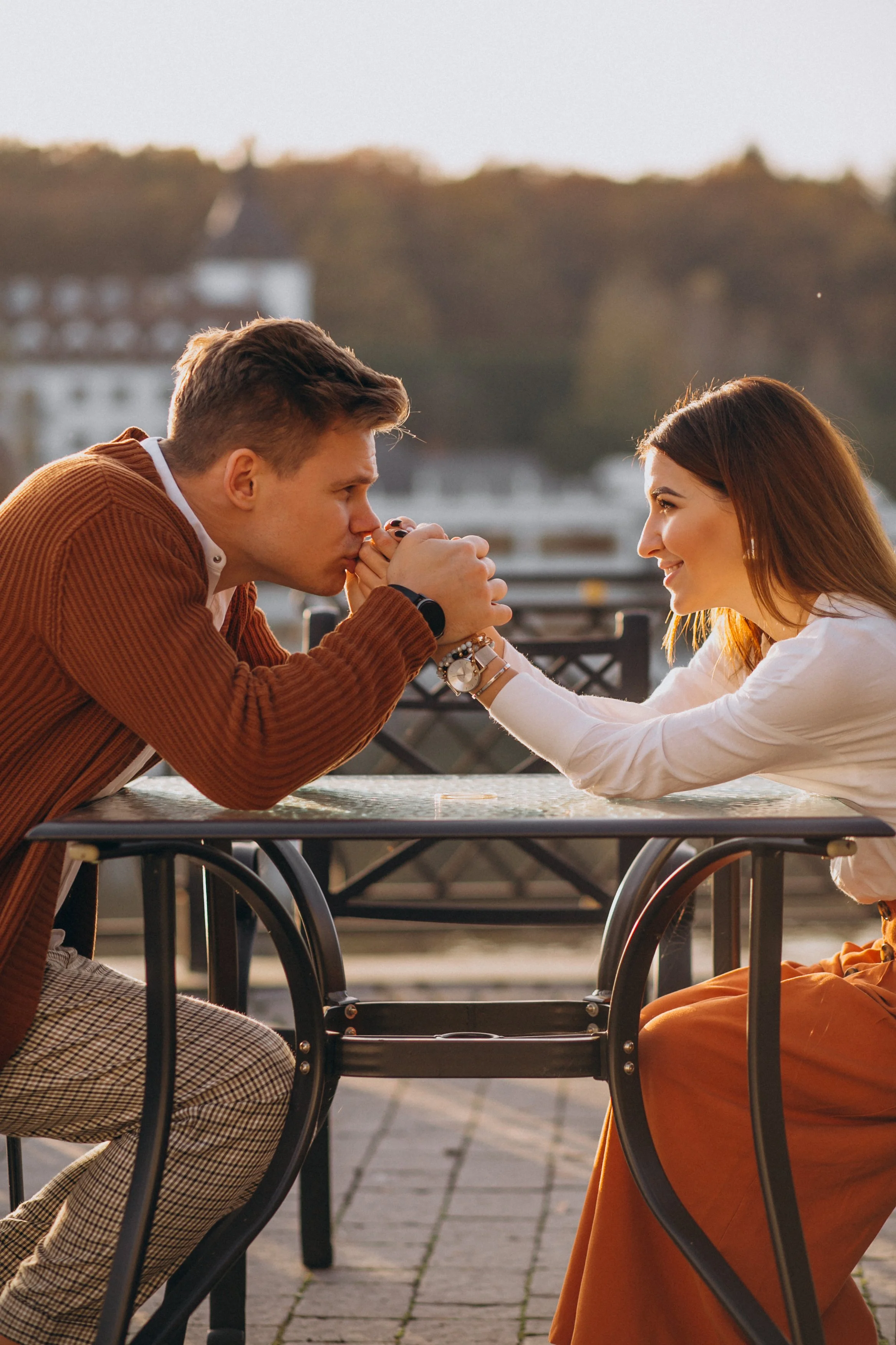 A young man and woman at an outdoor table, holding hands and smiling at each other during sunset.