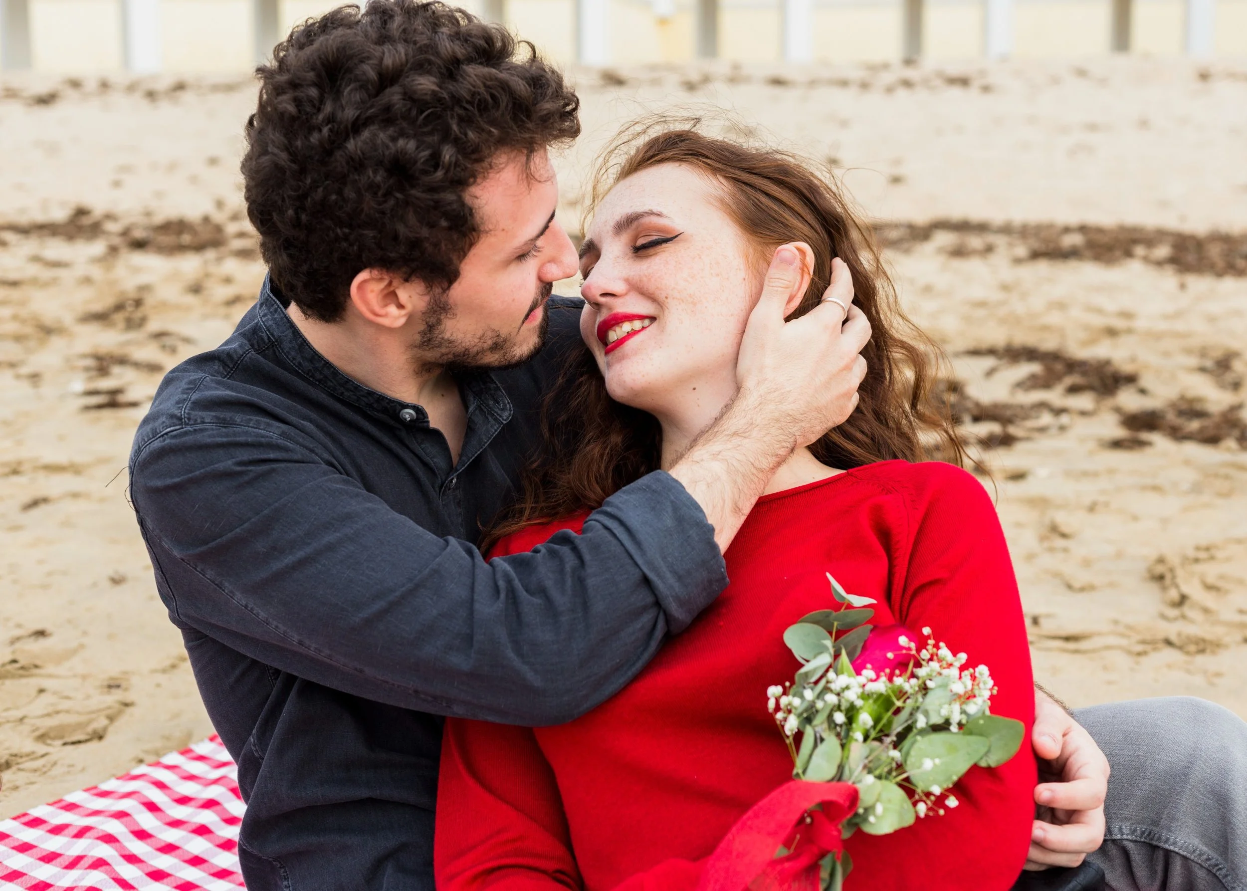 A couple sitting on a beach with the man holding the woman's face, both smiling and close to each other, with a bouquet of flowers in the woman's hand.