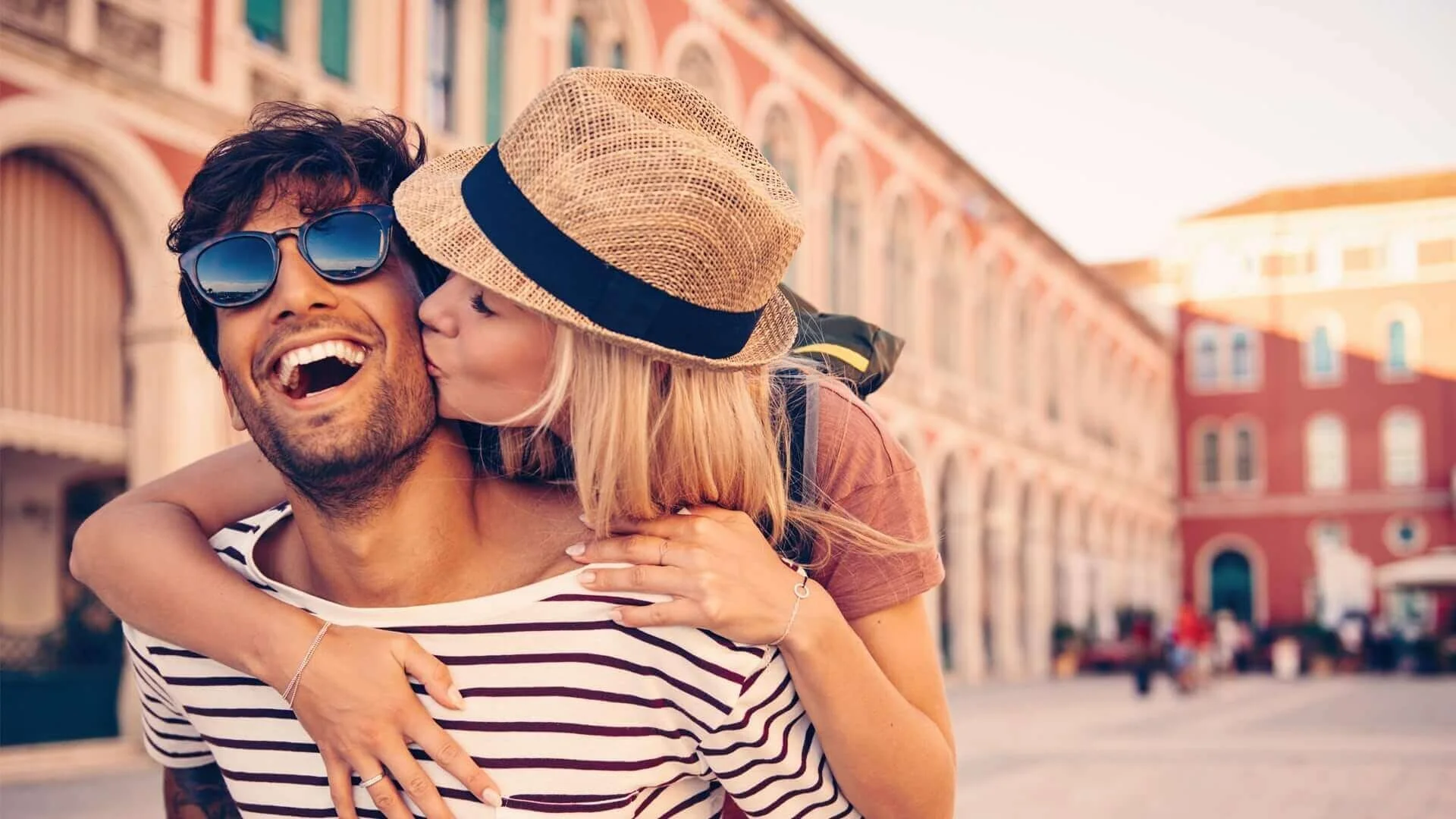 A couple enjoying a city walk, with the woman kissing the man on the cheek in front of a historic building.