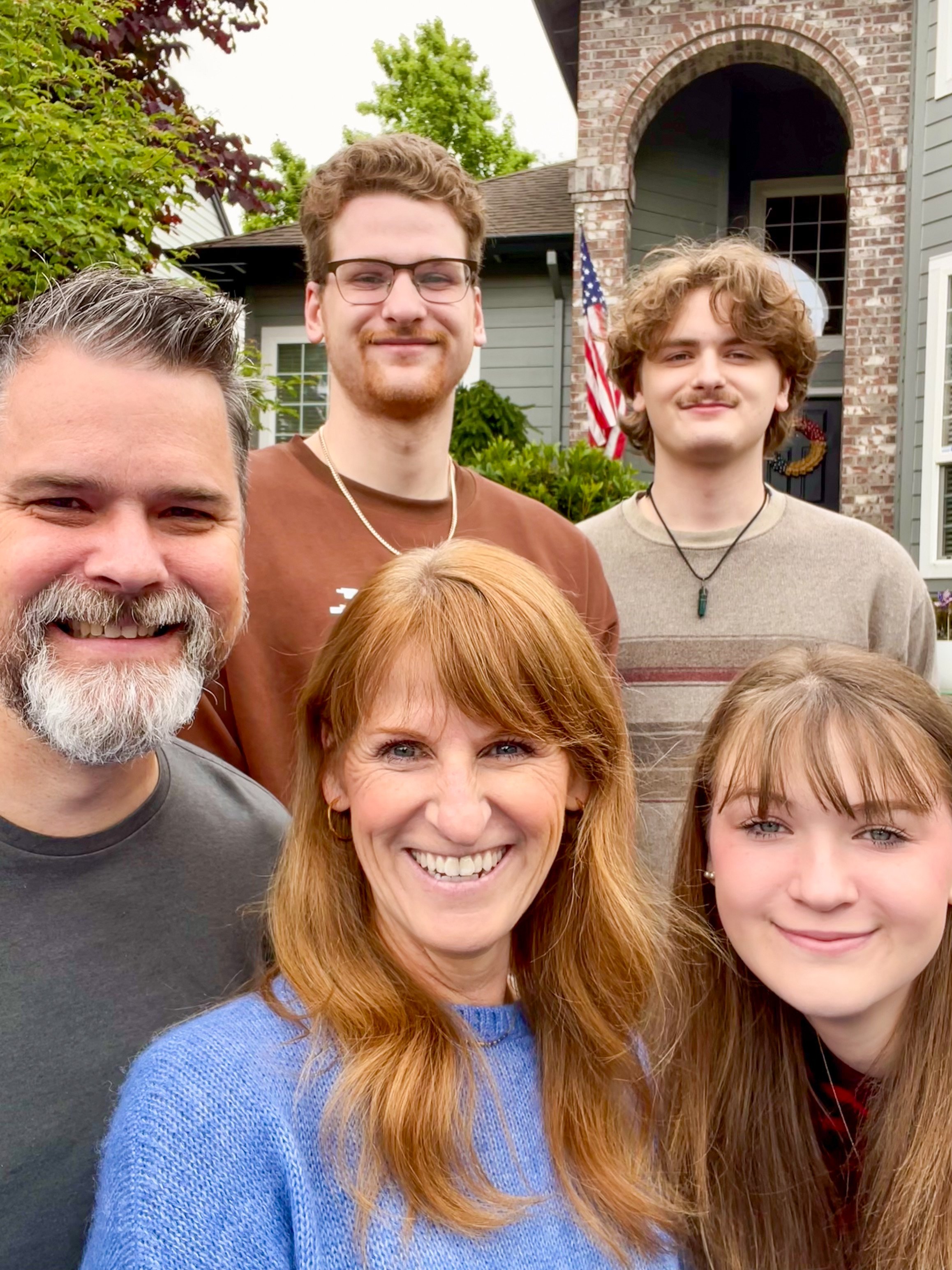 A family of six posing for a photo in front of their house, which has a brick arch and an American flag, with trees and greenery around.
