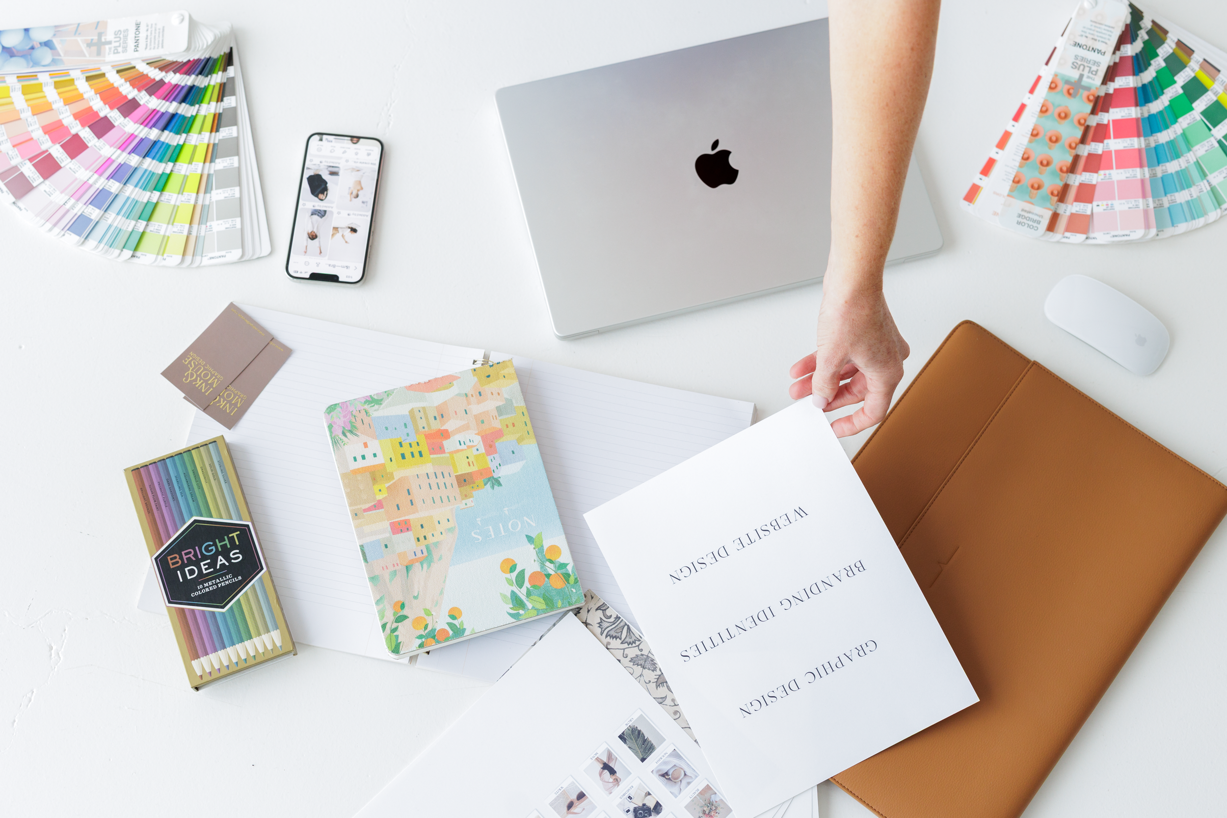 Design workspace with color swatches, patterned notebooks, iPhone displaying product images, MacBook, mouse, papers, and a brown portfolio on a white desk.