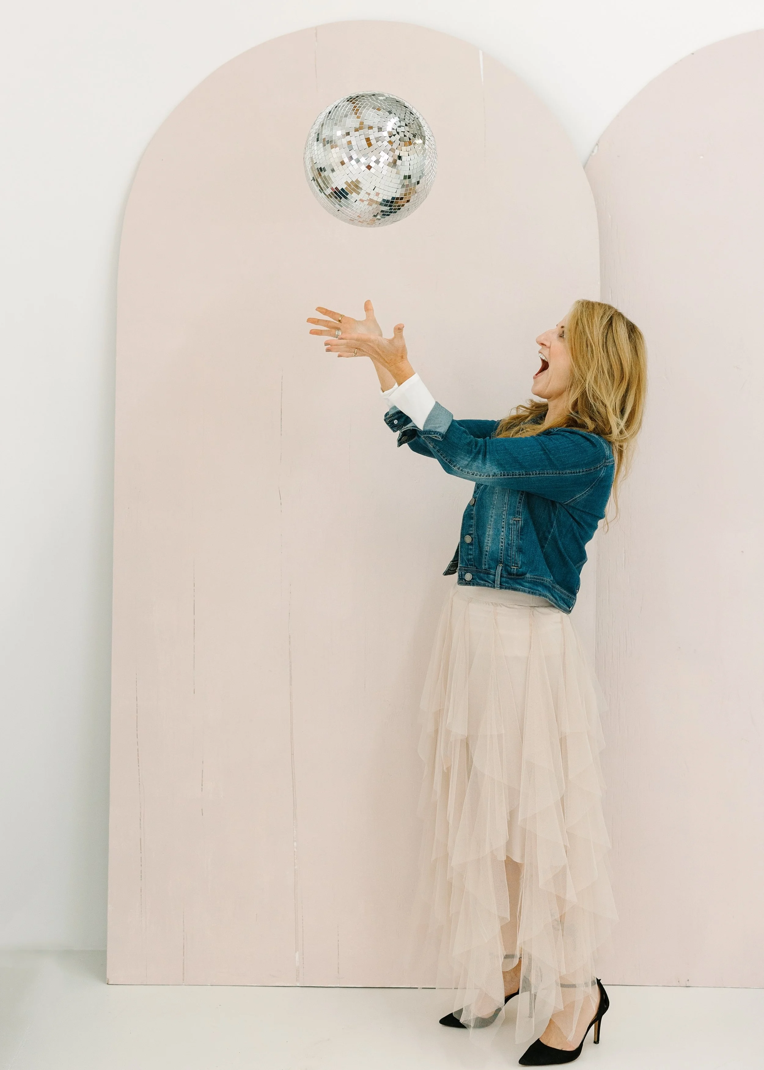 A woman in a denim jacket and beige tulle skirt reaches out towards a disco ball hanging from the ceiling in a bright room with light pink and white walls.