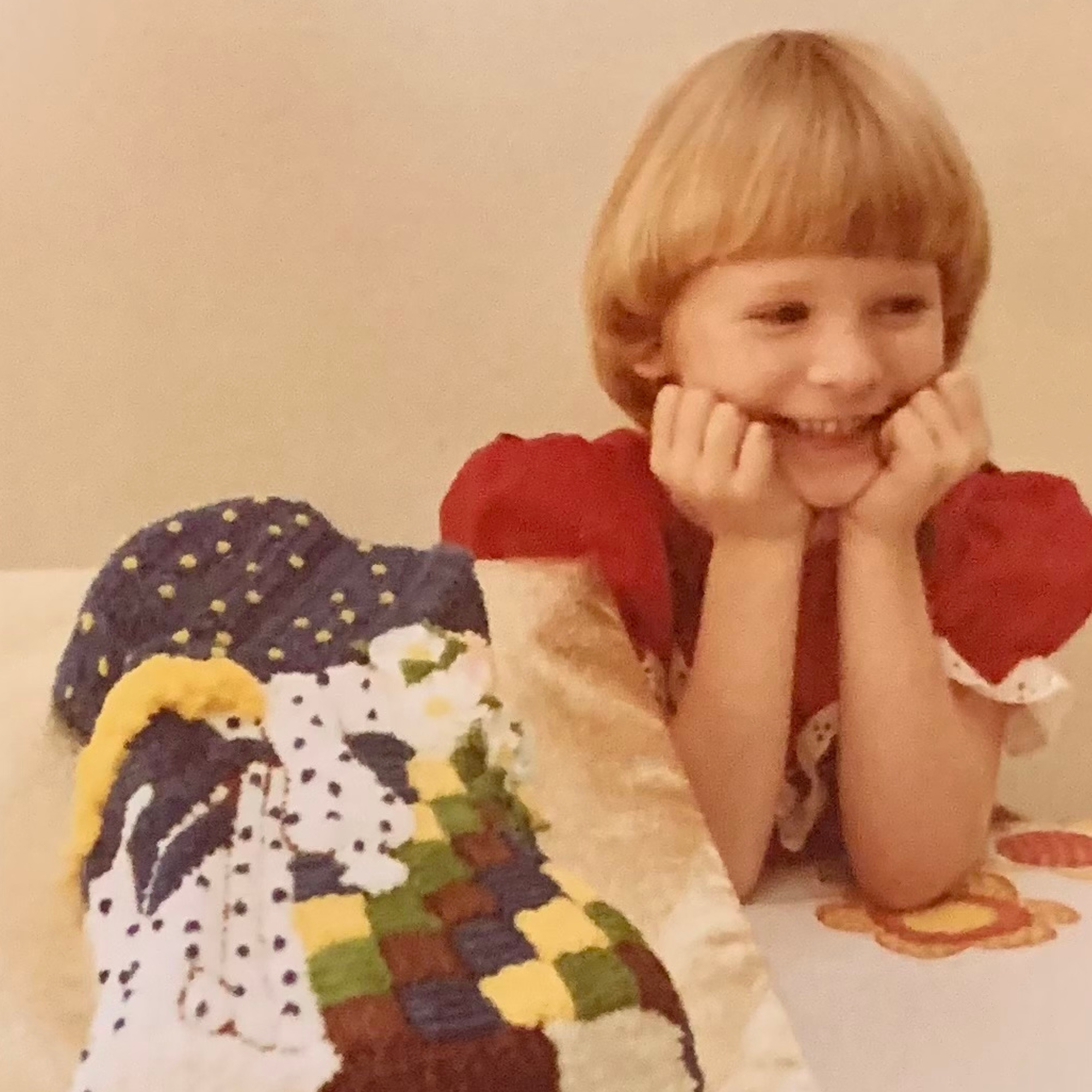 A young girl with short red hair and a red shirt, smiling and resting her chin on her hands, lying on a bed with a colorful patchwork quilt.