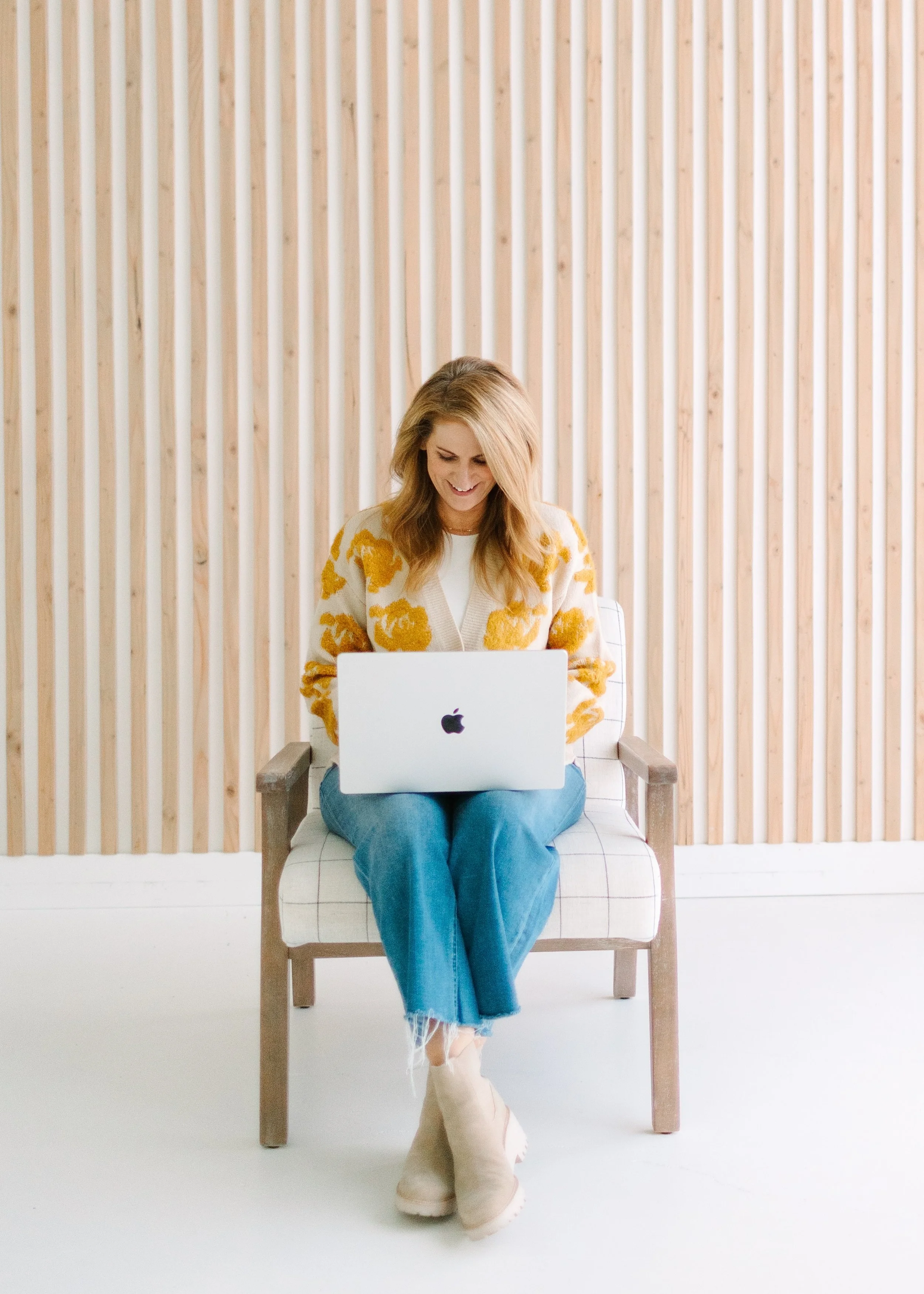 A woman with long red hair, wearing a white and orange sweater and blue jeans, sitting on a wooden chair with a white cushion, working on a silver MacBook in a room with a light wooden slat wall.